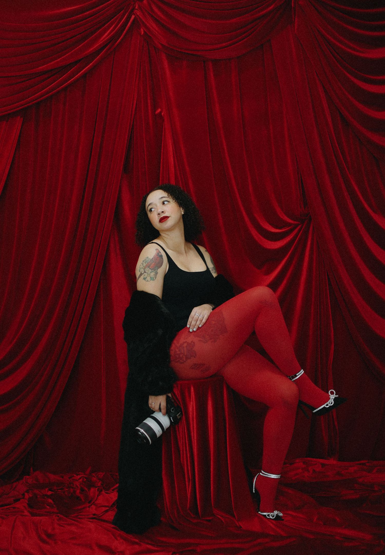 Female photographer in black top and red tights sitting with camera against red velvet backdrop.