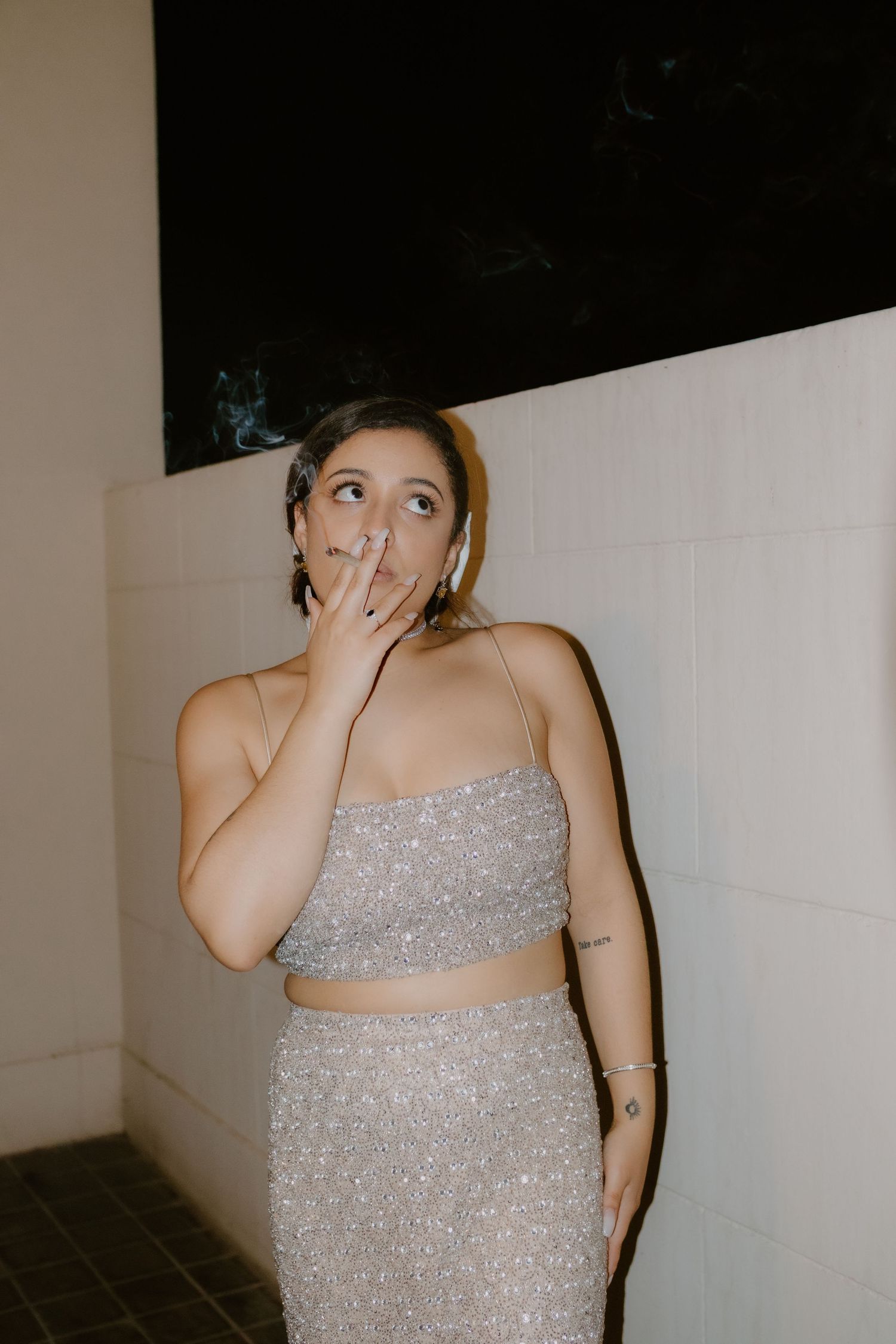 Young woman in silver sequin two-piece outfit smoking a cigarette against white tiled wall at wedding reception.