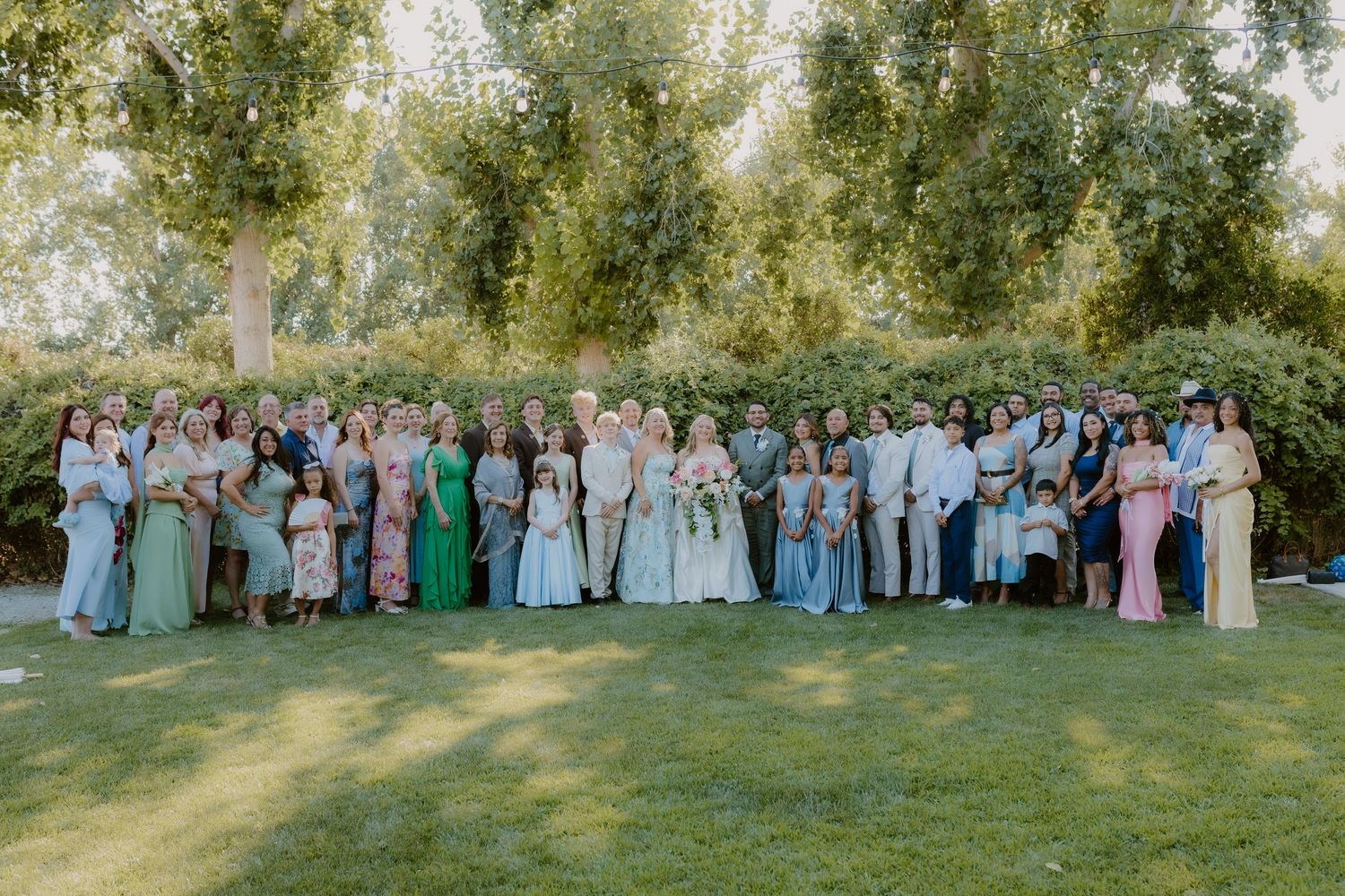 Large wedding group portrait with bride and groom surrounded by guests on green lawn.