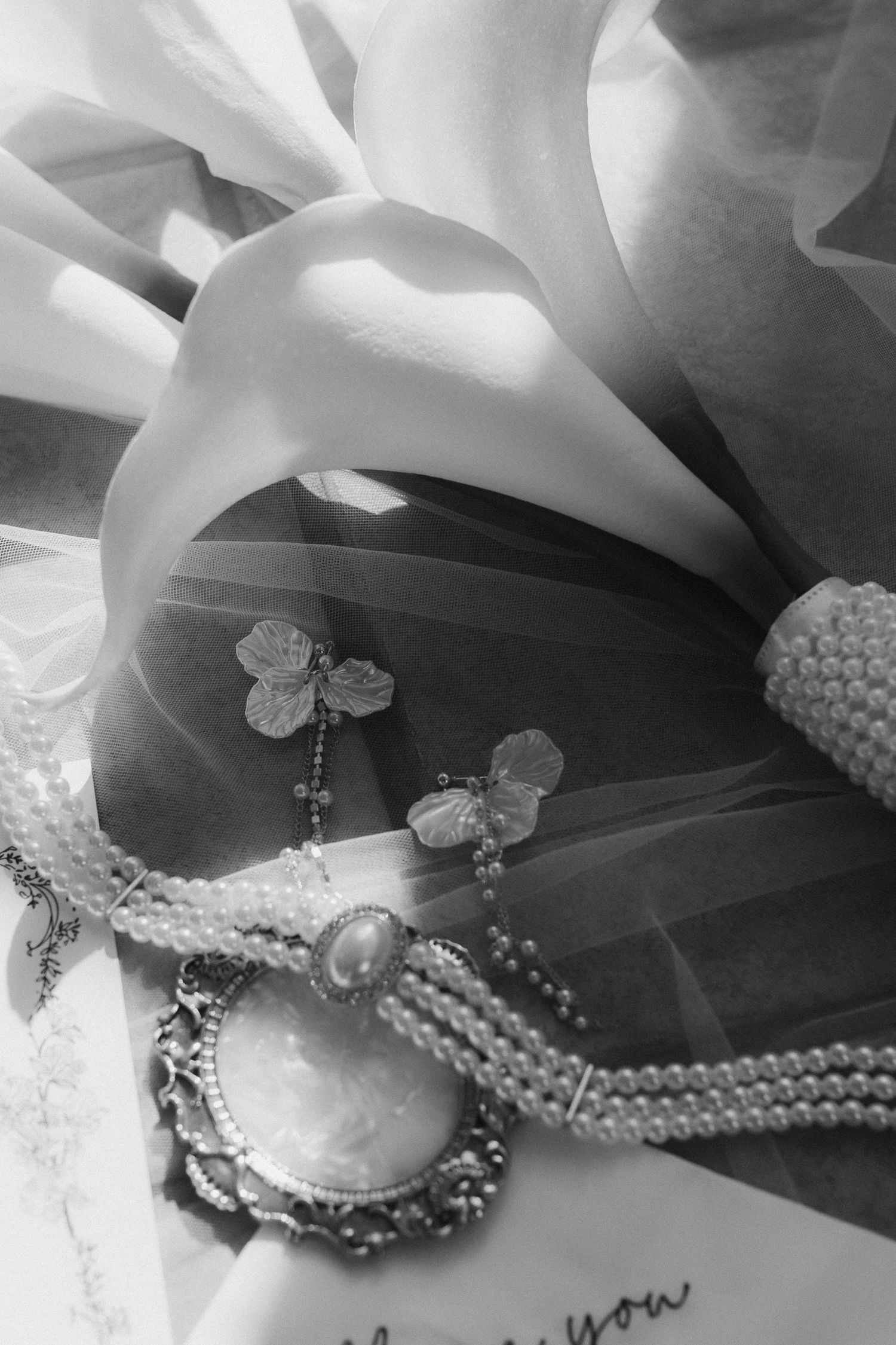 Black and white flat lay of bridal jewelry including pearl necklace, cameo brooch, and butterfly earrings on white fabric.