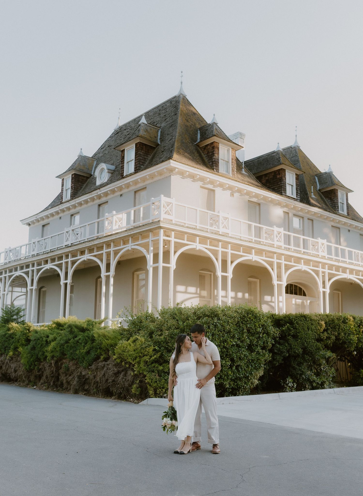 Couple in white attire sharing a kiss in front of a stunning multi-story Victorian mansion at dusk.