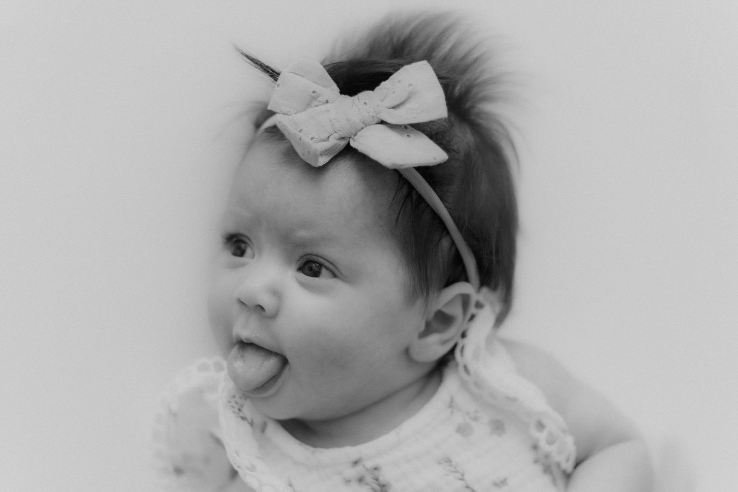 Black and white portrait of cute baby girl with bow headband sticking tongue out.