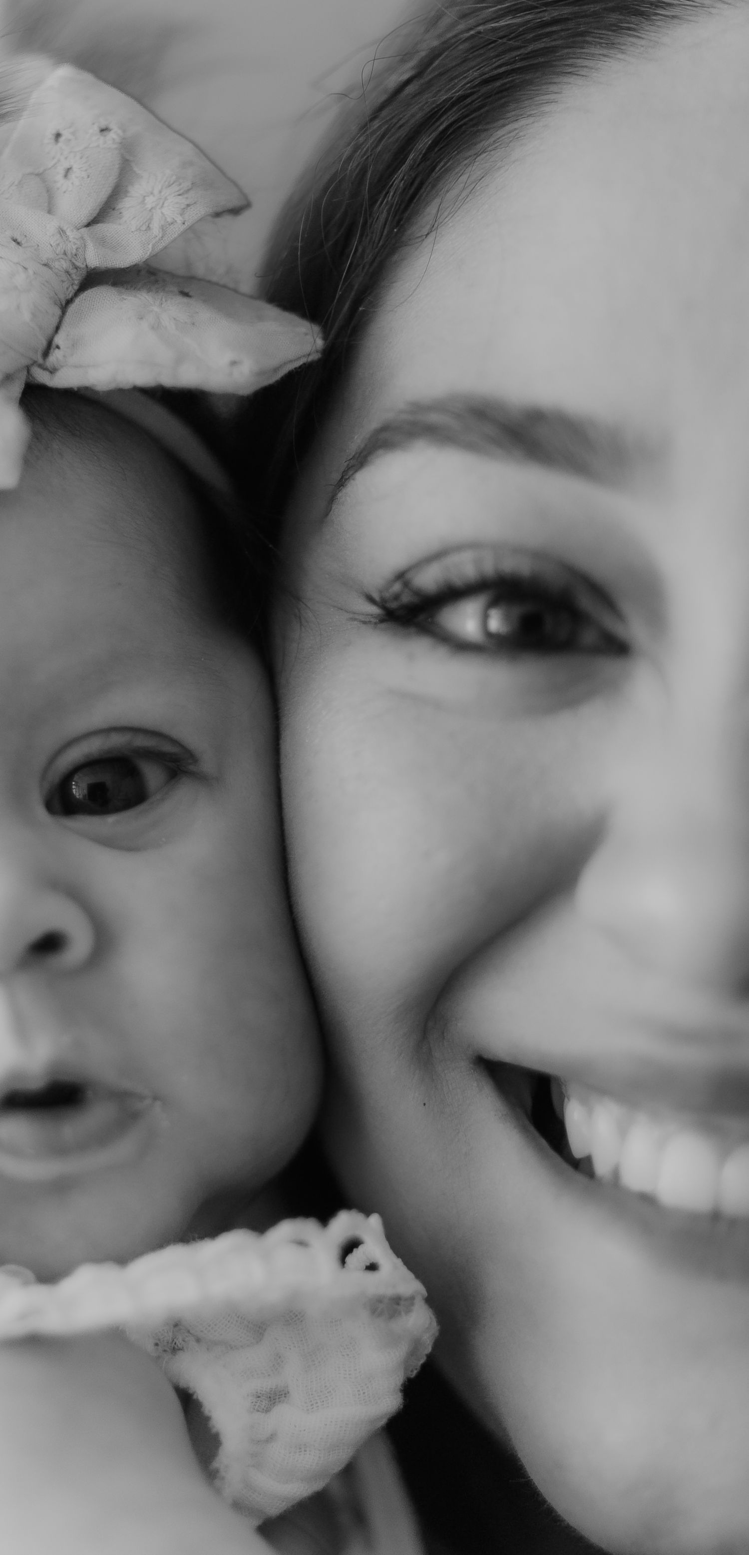 Black and white close-up portrait of a smiling mother cheek-to-cheek with her baby girl wearing a bow headband.
