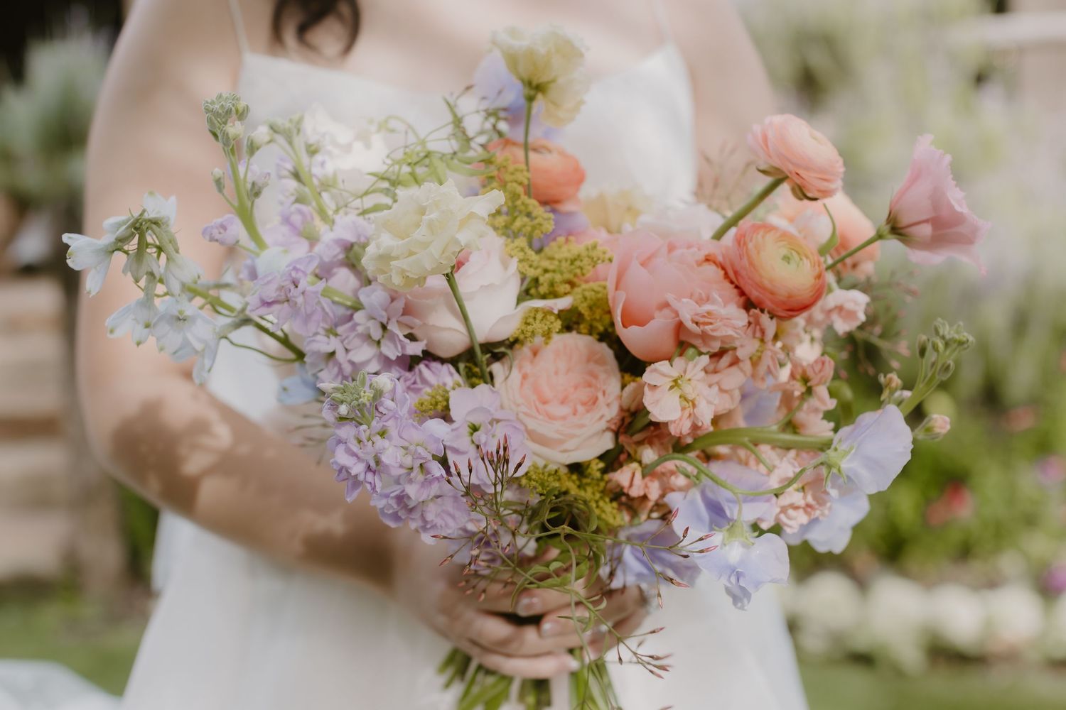 Bride holding a romantic pastel wedding bouquet with pink ranunculus, lavender, and peach blooms.