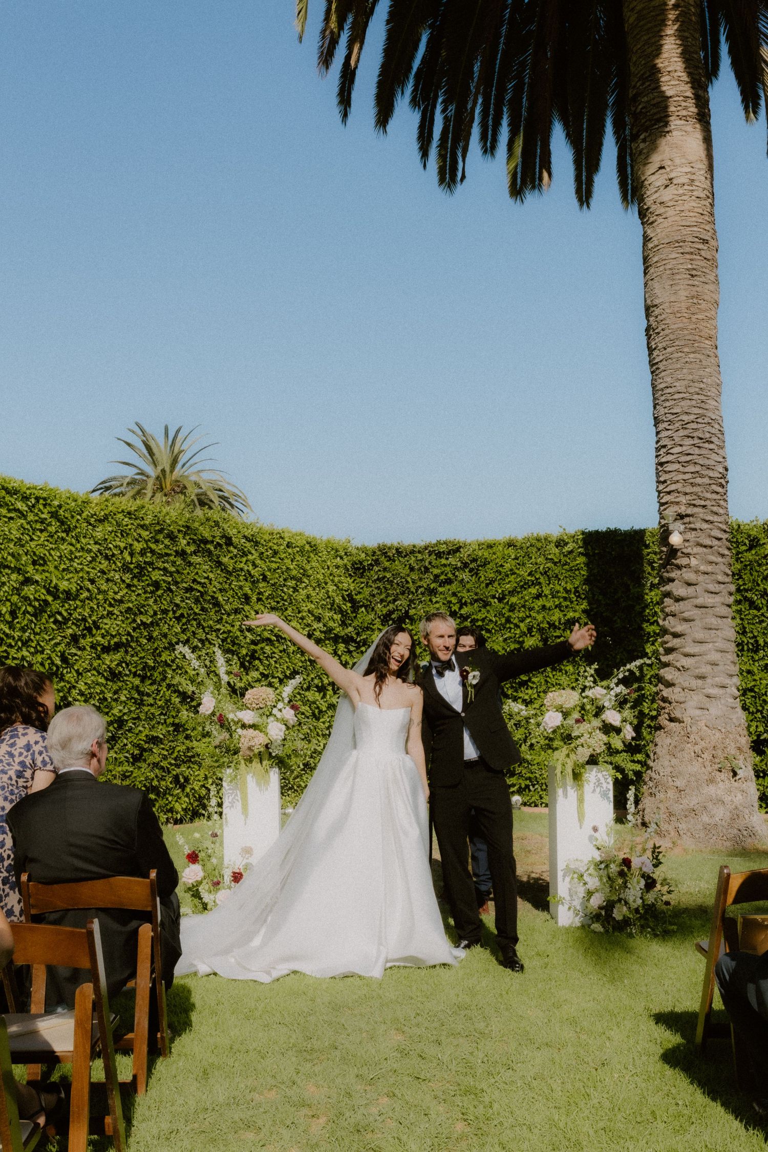 Newlyweds celebrate recessional on grass aisle surrounded by guests at outdoor garden wedding ceremony.