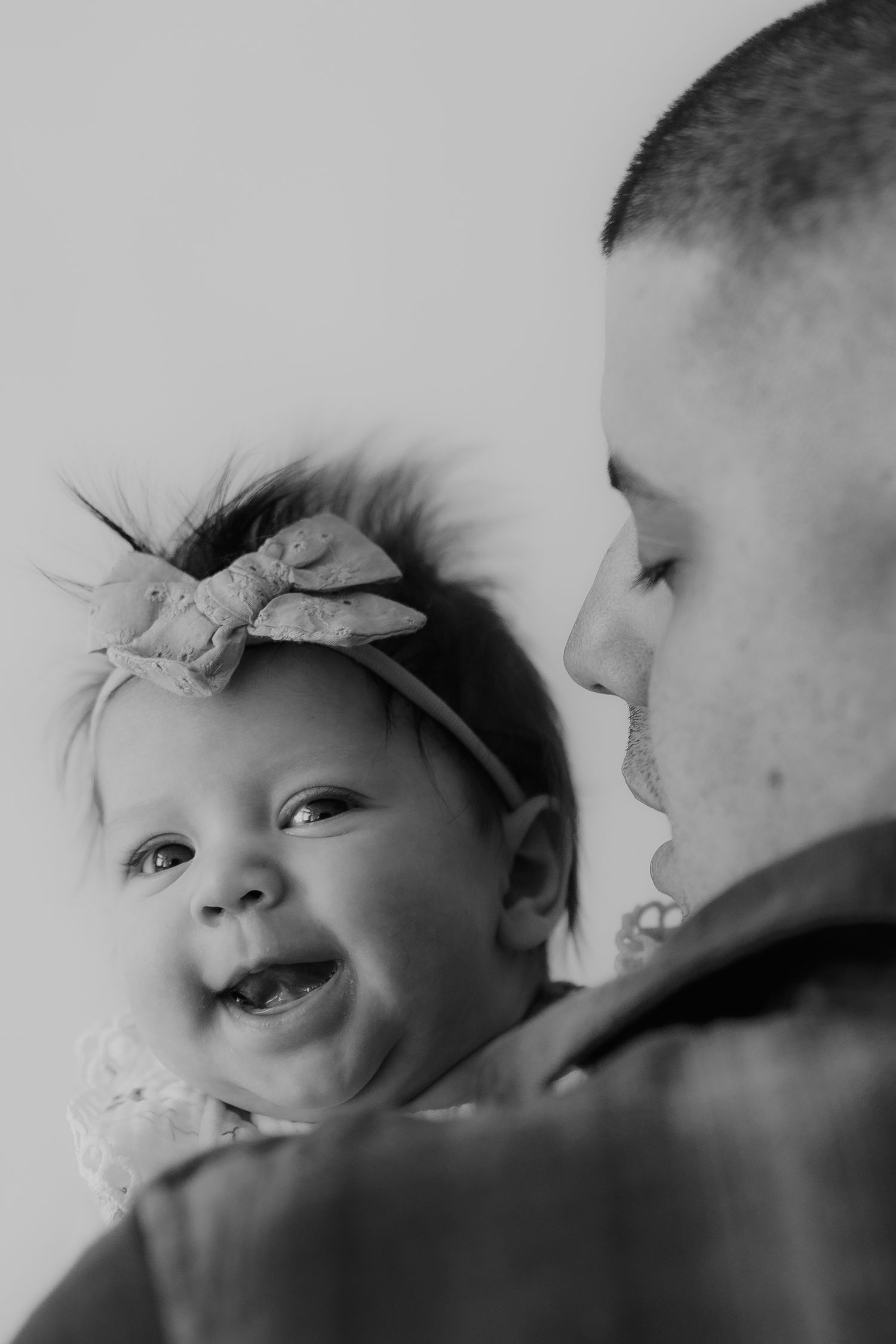 Black and white photo of a laughing newborn baby girl with bow being held by her smiling father.