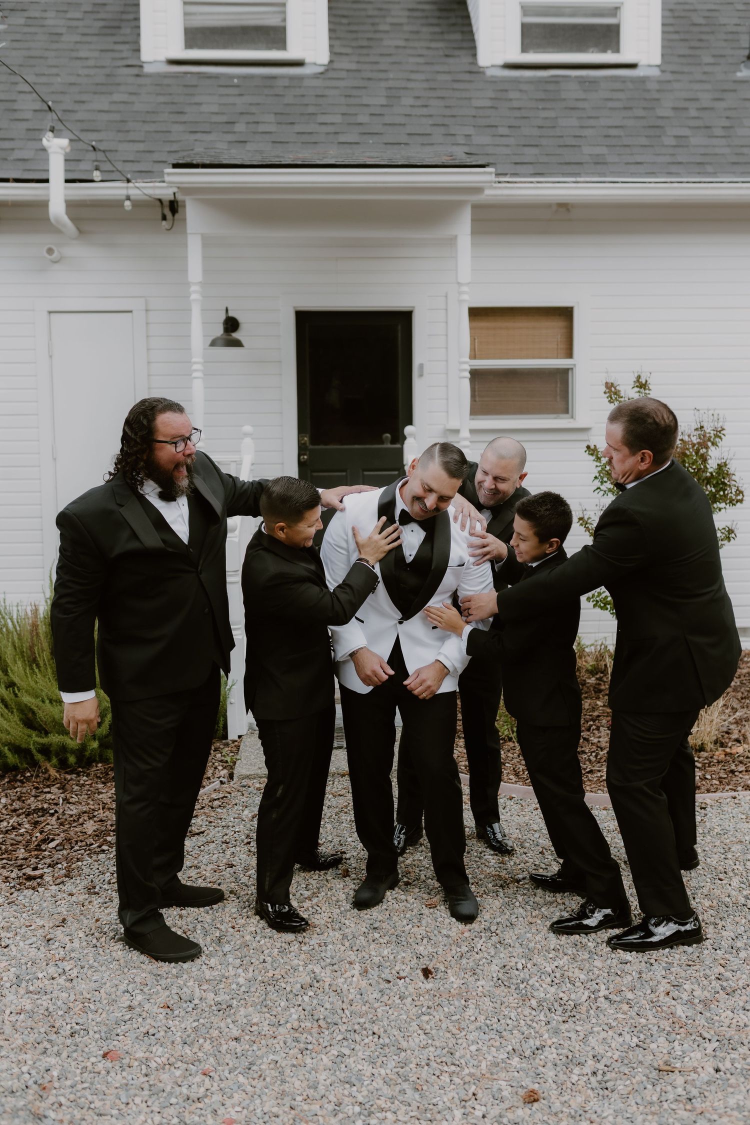 Groom in white jacket surrounded by groomsmen in black tuxedos sharing a joyful moment outside white building.
