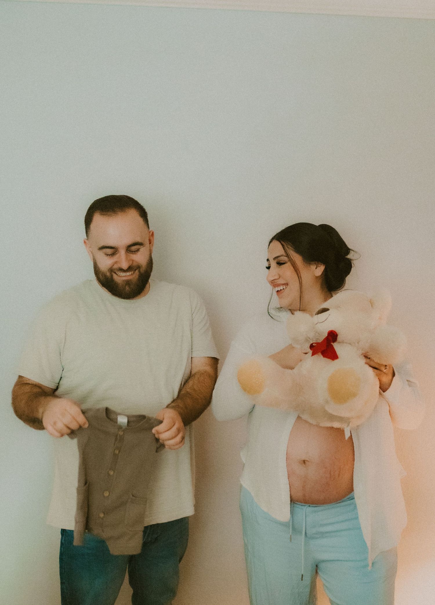 Smiling couple holding tiny baby clothes and teddy bear during a maternity photo session indoors.