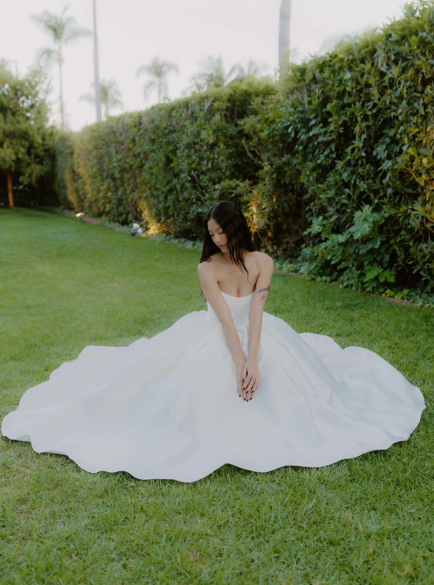 Bride in a stunning white ballgown wedding dress sitting on green grass with lush hedges backdrop.