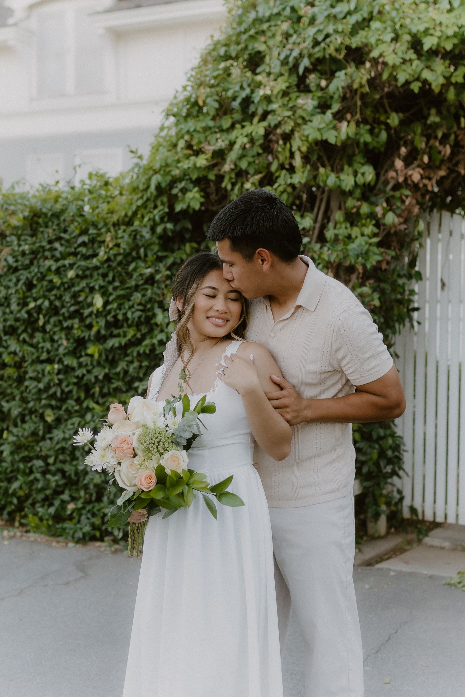 Groom kissing bride on the cheek as she smiles holding a pastel floral bouquet outdoors.