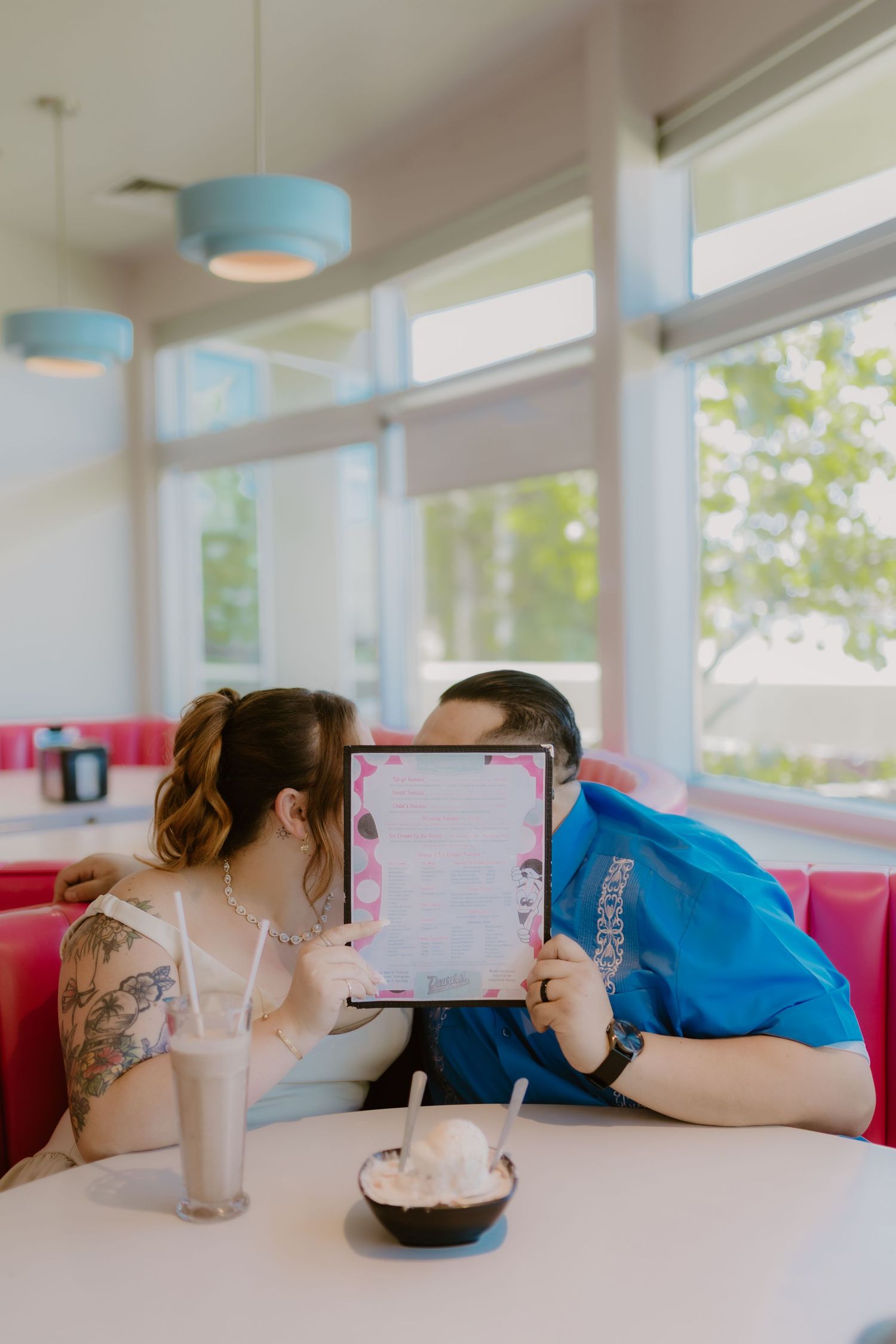 Couple kissing behind pink diner menu at retro booth with milkshakes and ice cream on table. Bakersfield Photographer