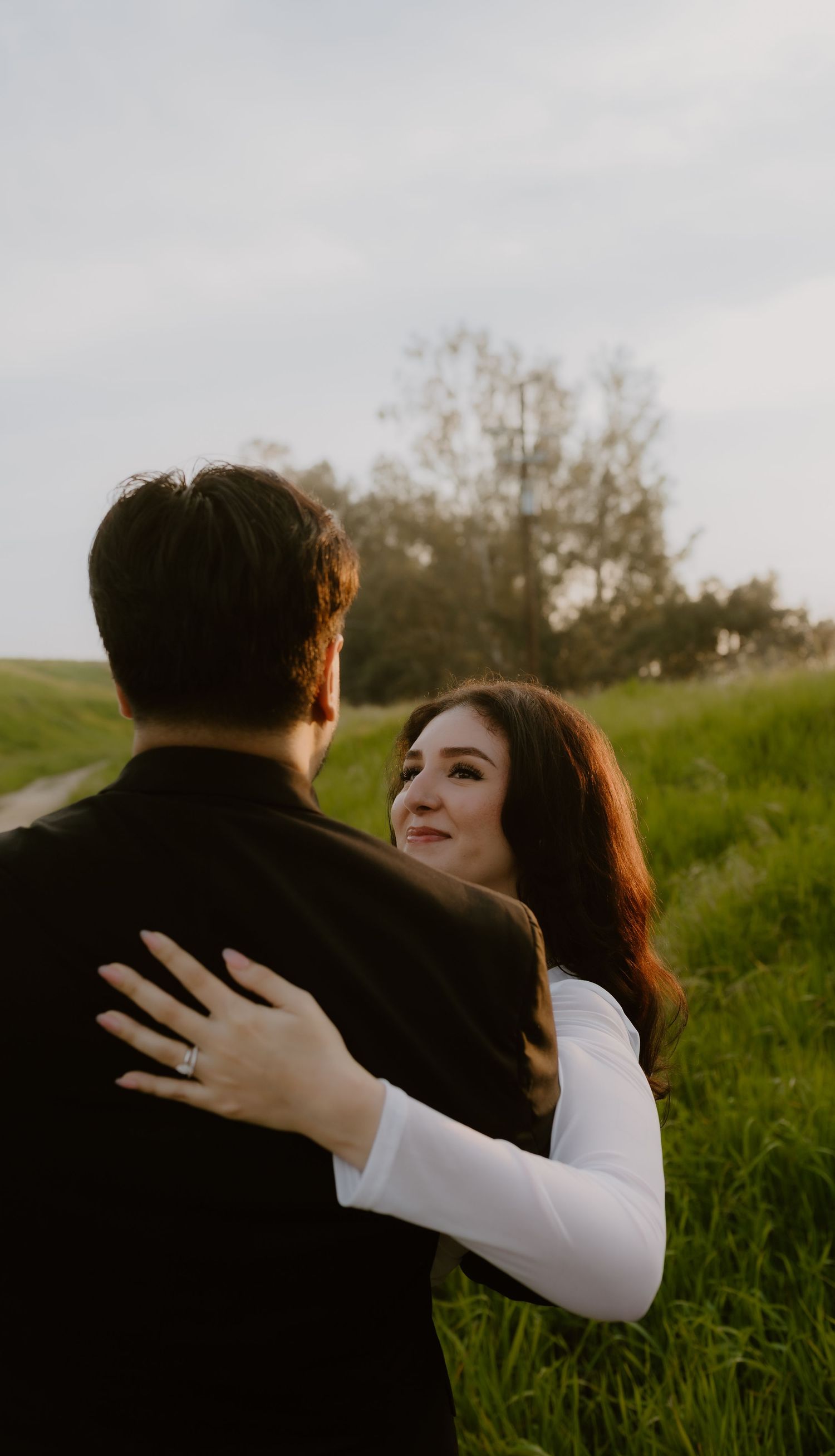 A woman with auburn hair embraces her partner from behind while gazing up at him in a green field.