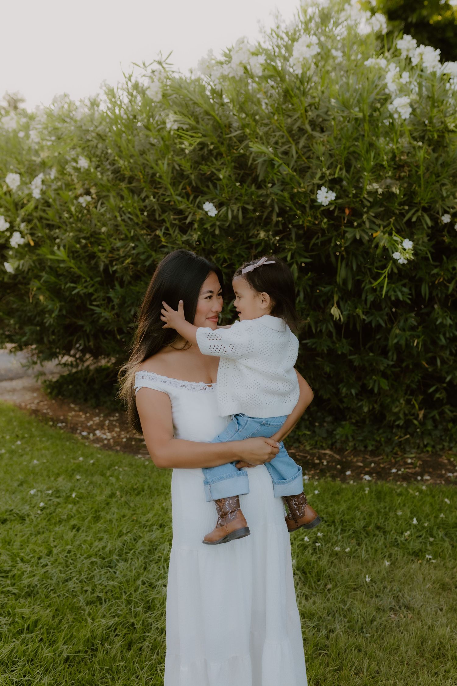 Mother in white off-shoulder dress lovingly holding her toddler daughter in a garden setting.