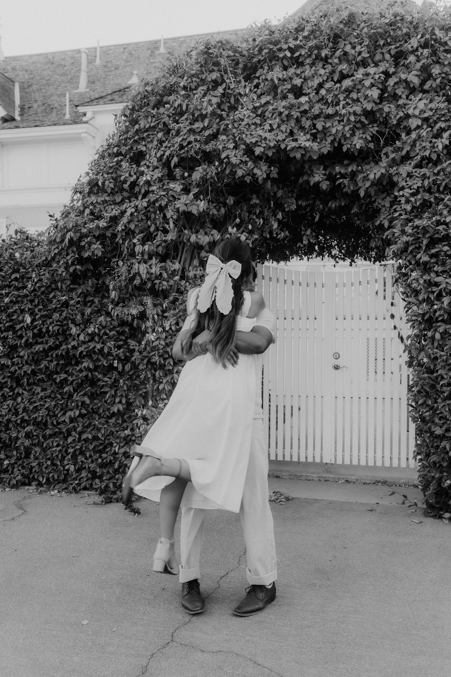 Black and white photo of couple embracing near ivy-covered white gate, woman wearing bow.