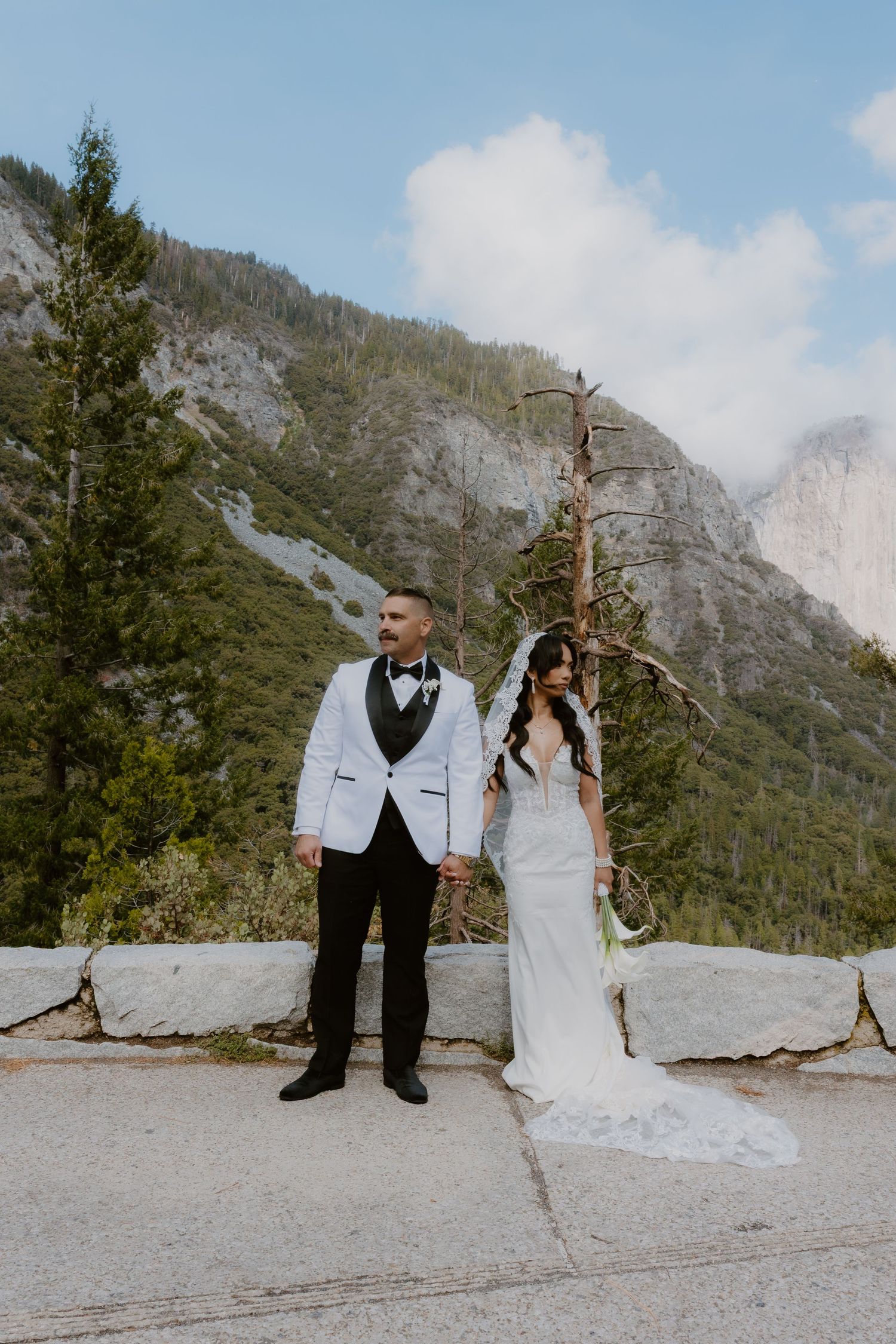 Bride and groom hold hands at Yosemite Valley overlook, groom in white tuxedo jacket, bride in lace gown.