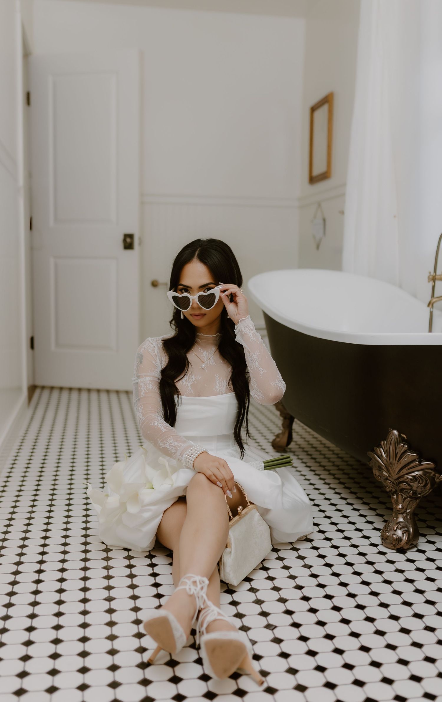 Bride in lace wedding dress sits on black and white tile floor, wearing heart-shaped sunglasses.