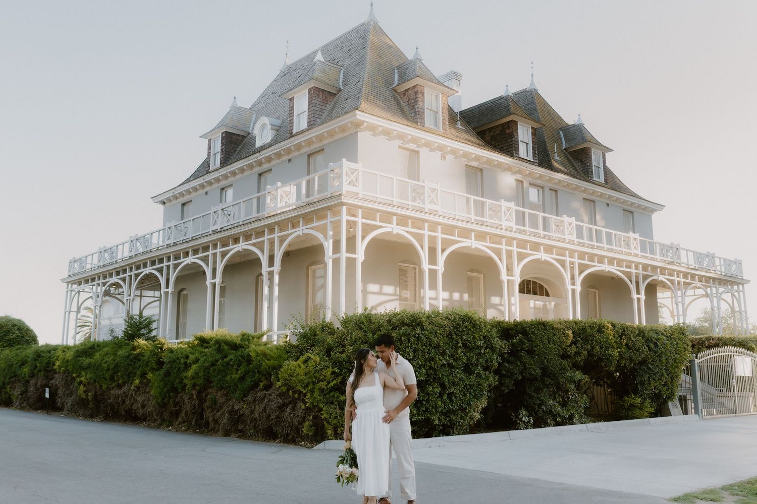 Couple sharing a romantic kiss in front of a grand white Victorian mansion during their engagement shoot.