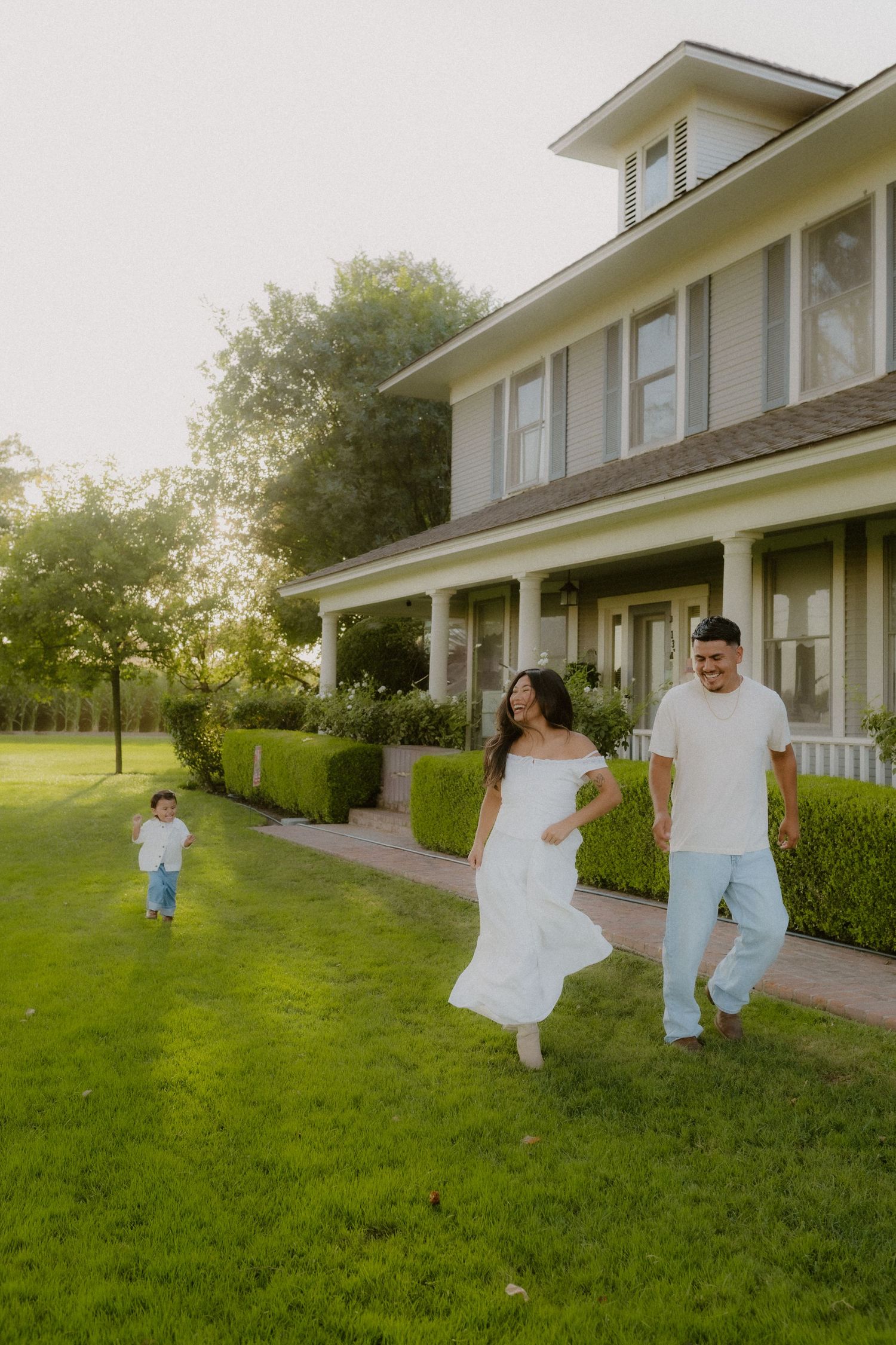 Happy family in white outfits walking on lush green lawn in front of large farmhouse at golden hour.