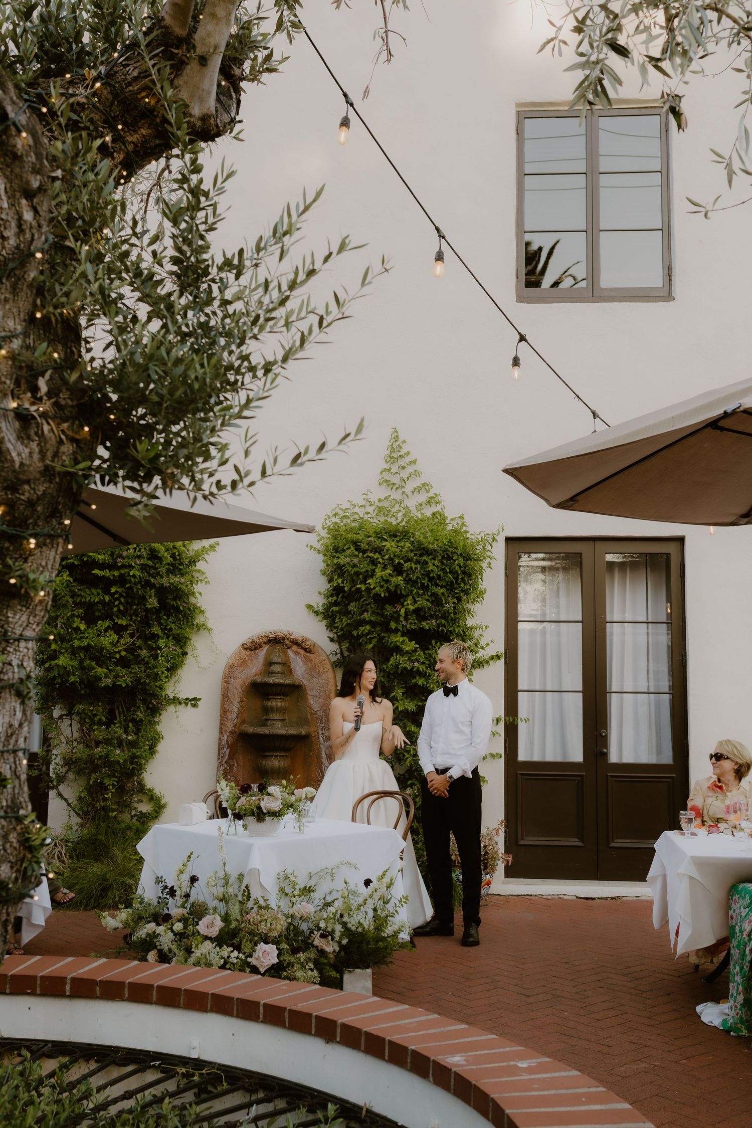 Wedding couple stands at their sweetheart table in a charming white courtyard with string lights and lush greenery.