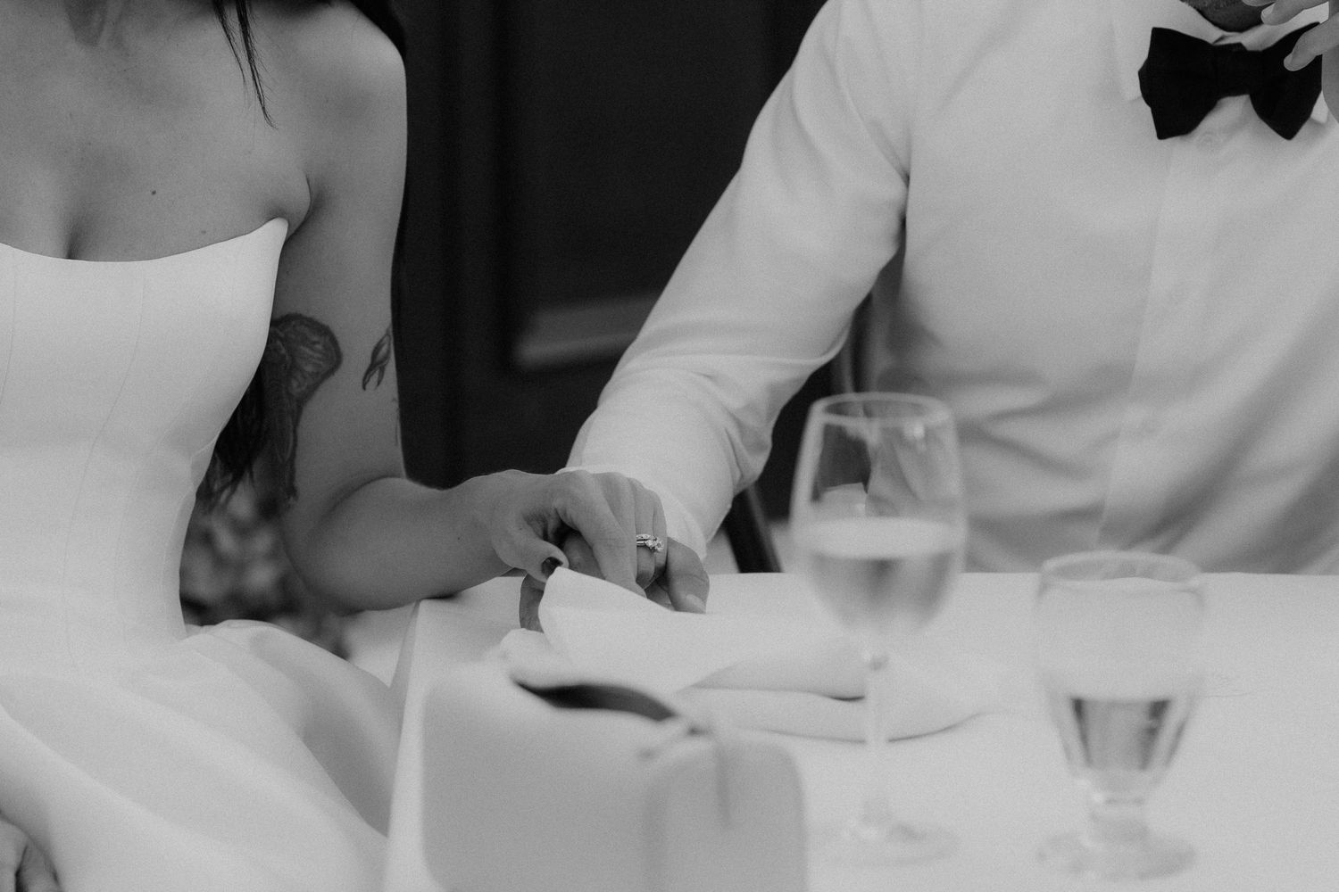 Black and white photo of bride and groom holding hands at a reception table with wine glasses nearby.