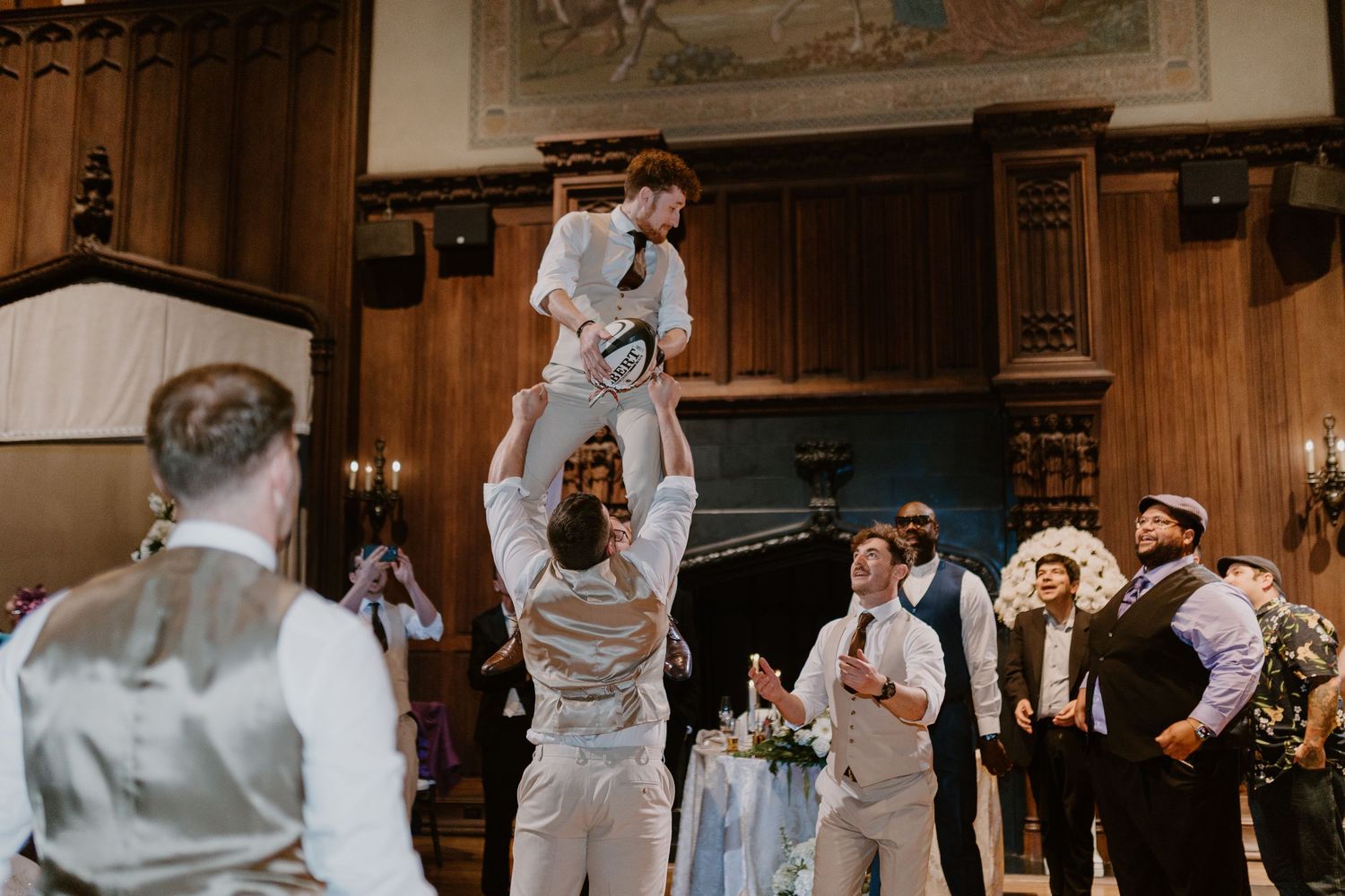 Groomsmen celebrating at a wedding reception in an elegant wood-paneled hall, lifting a child in the air.
