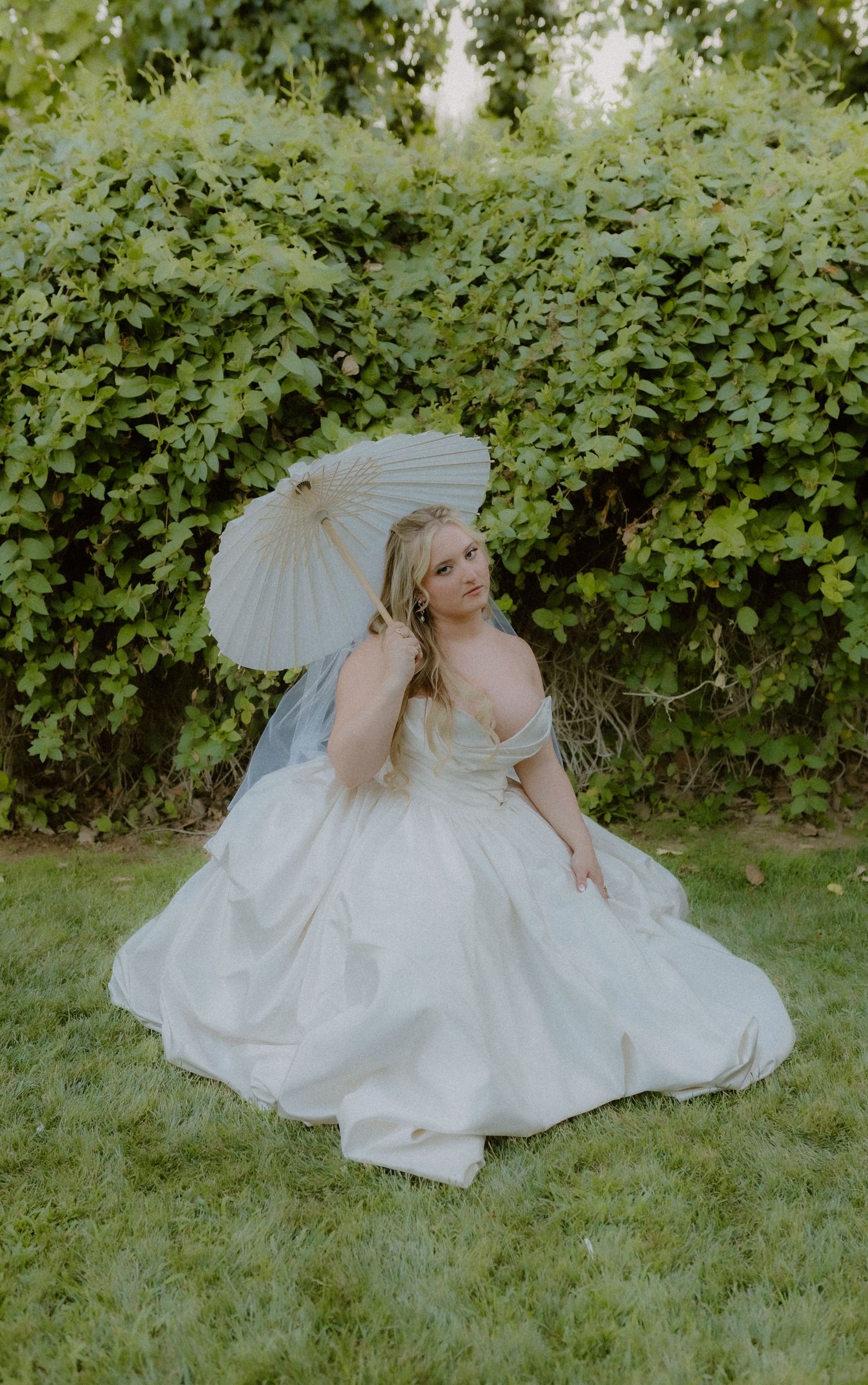 Elegant bride in a white ball gown sits on grass holding a parasol umbrella in front of a green hedge.