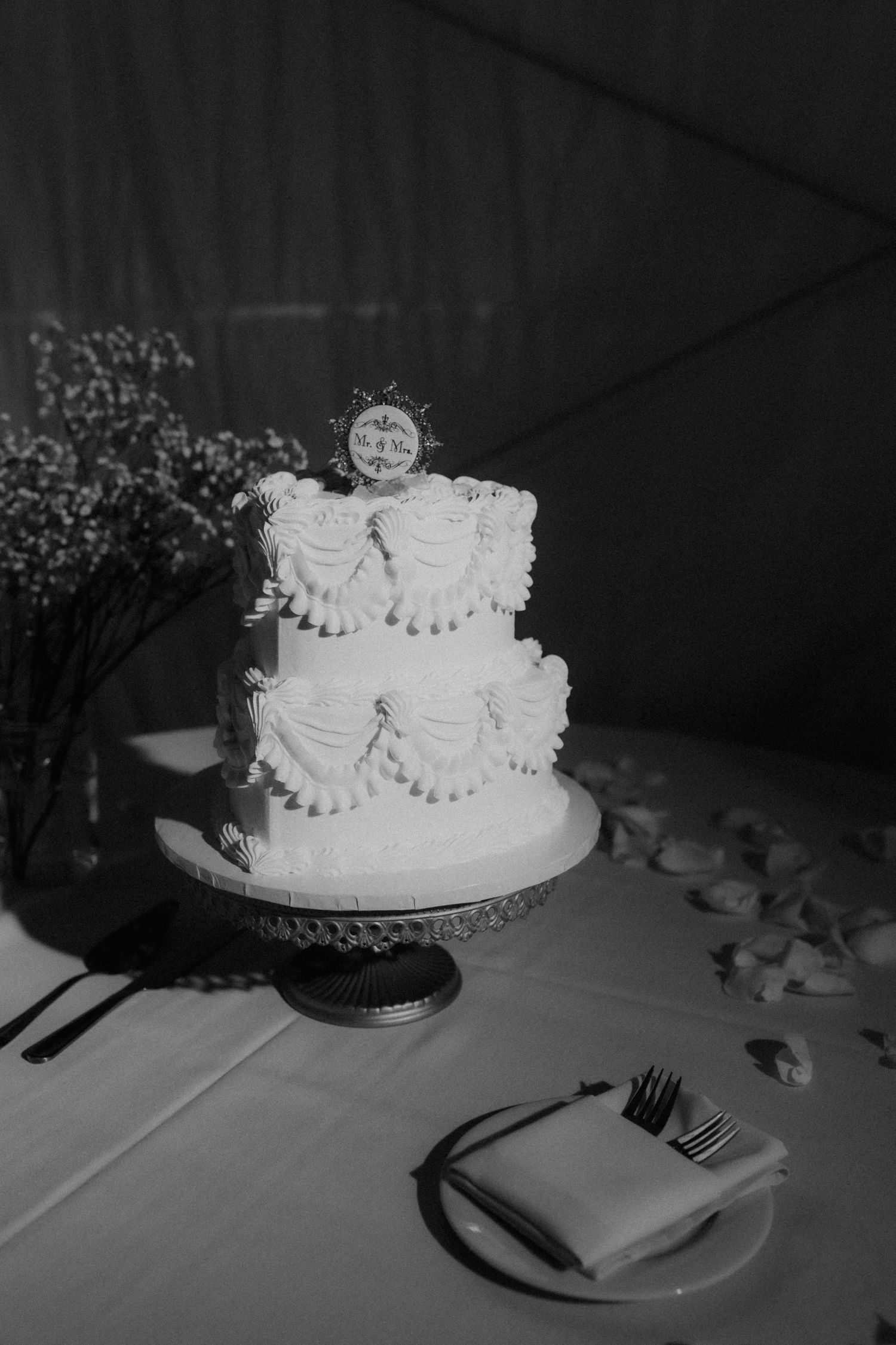Elegant two-tier white wedding cake with decorative frosting and sun topper on ornate cake stand.