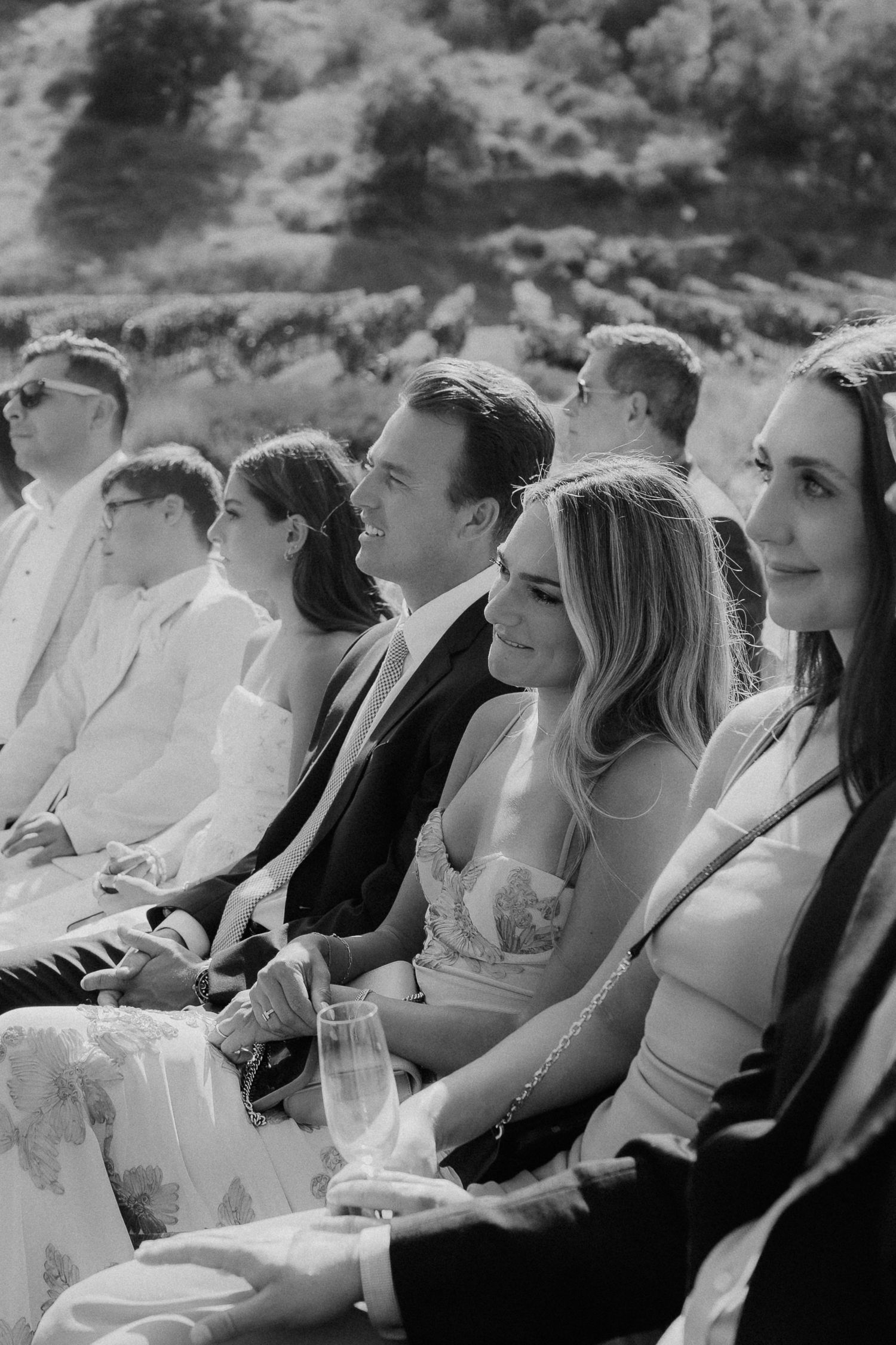 Black and white photo of smiling wedding guests seated together during an outdoor ceremony.