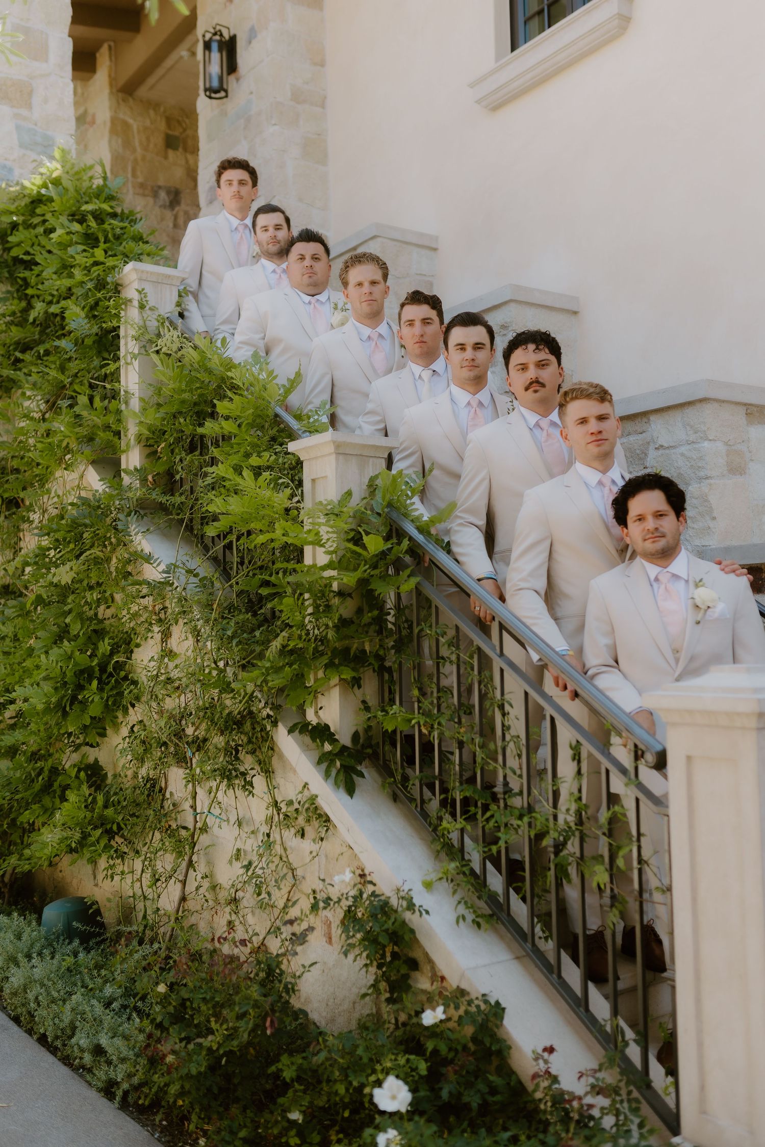 Groomsmen in tan suits pose on an outdoor staircase surrounded by lush greenery at a wedding.