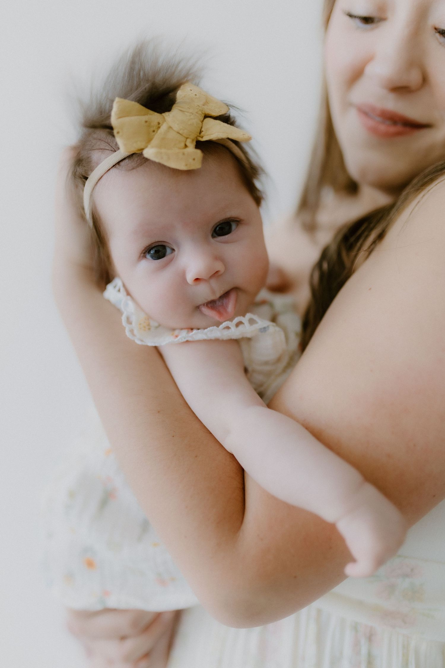 Adorable newborn baby girl with a gold bow headband sticking out her tongue while cradled in mothers arms.