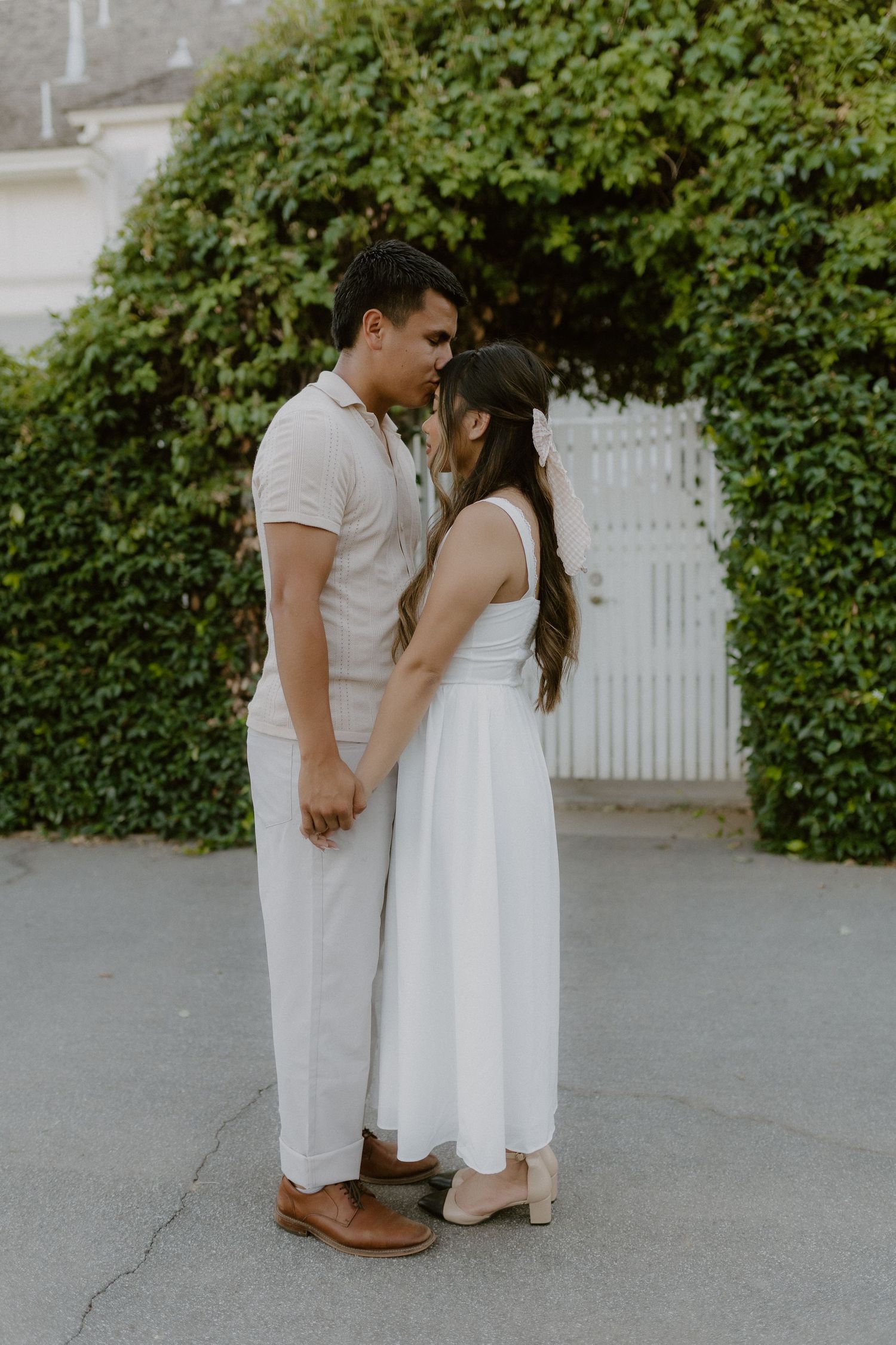 Couple in white outfits standing close together touching foreheads near ivy gate.