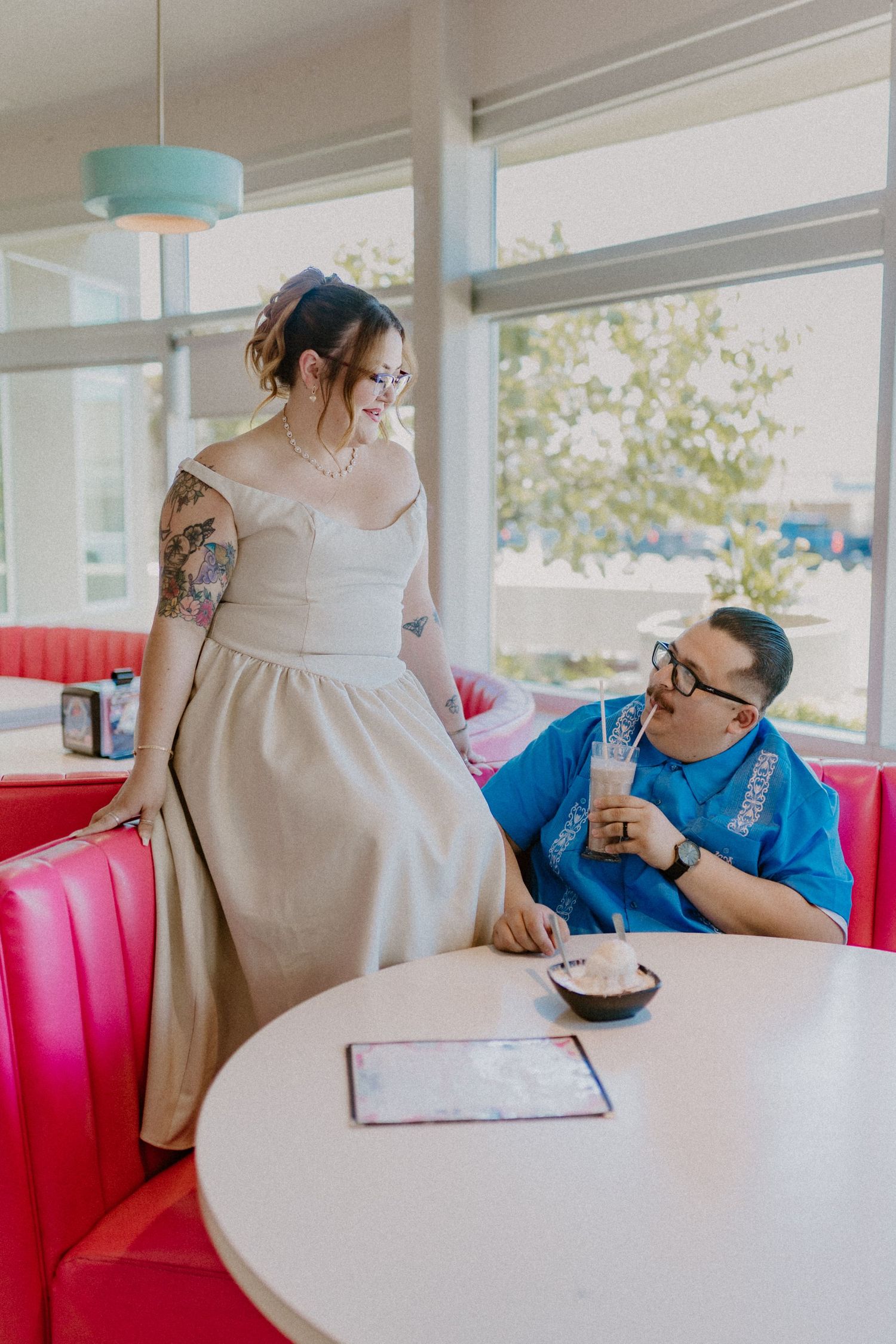 Bride in elegant off-shoulder gown standing beside groom sipping milkshake at pink retro diner booth.