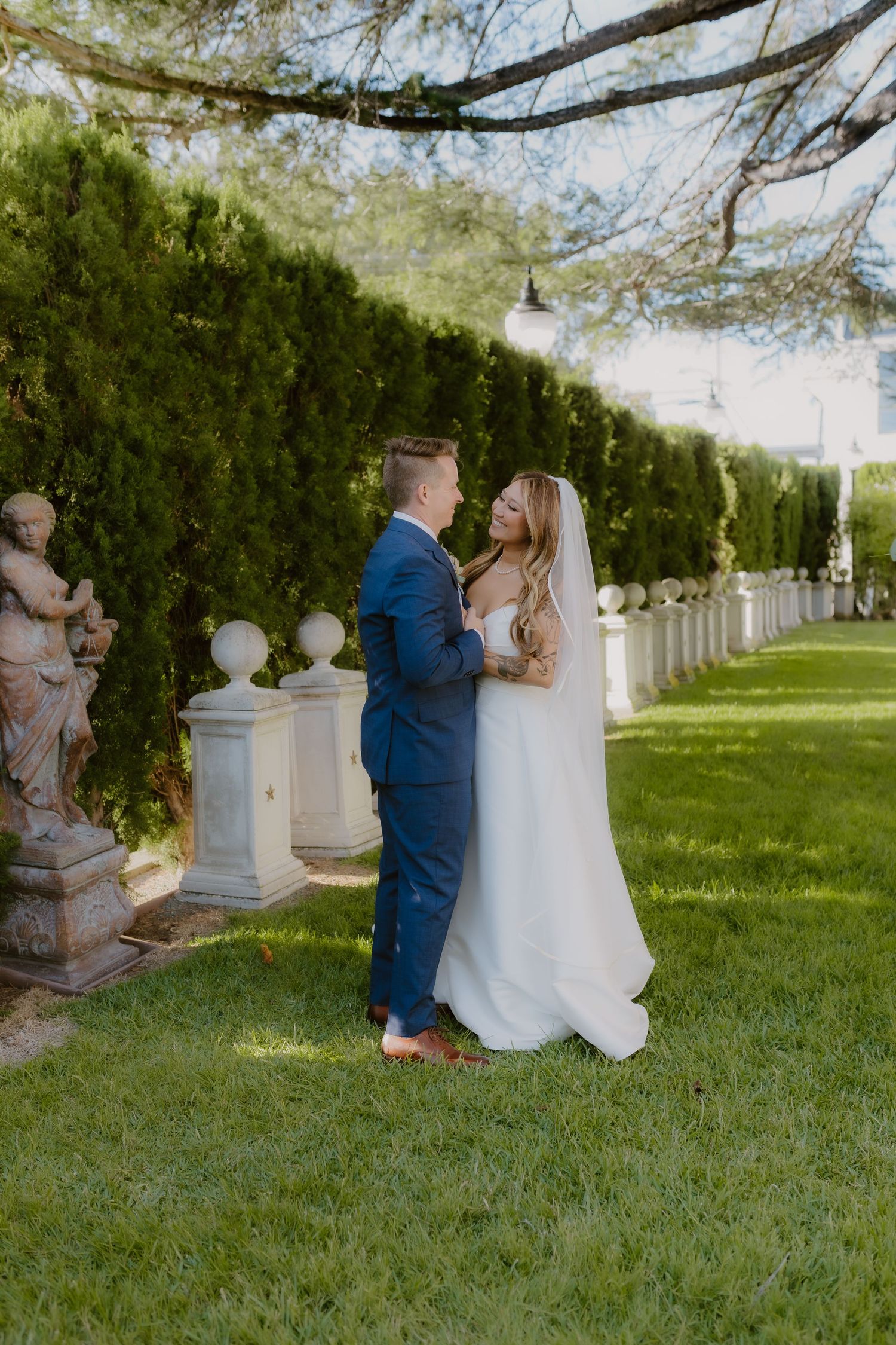 Bride and groom share a romantic moment in a lush garden with classical statues, groom in navy suit. Jefferson Street Mansion, North Photo