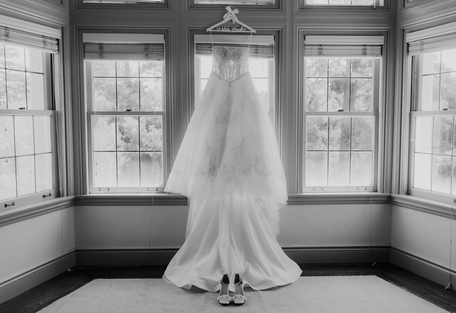 Black and white photo of wedding gown and veil hanging in window with bridal shoes below.