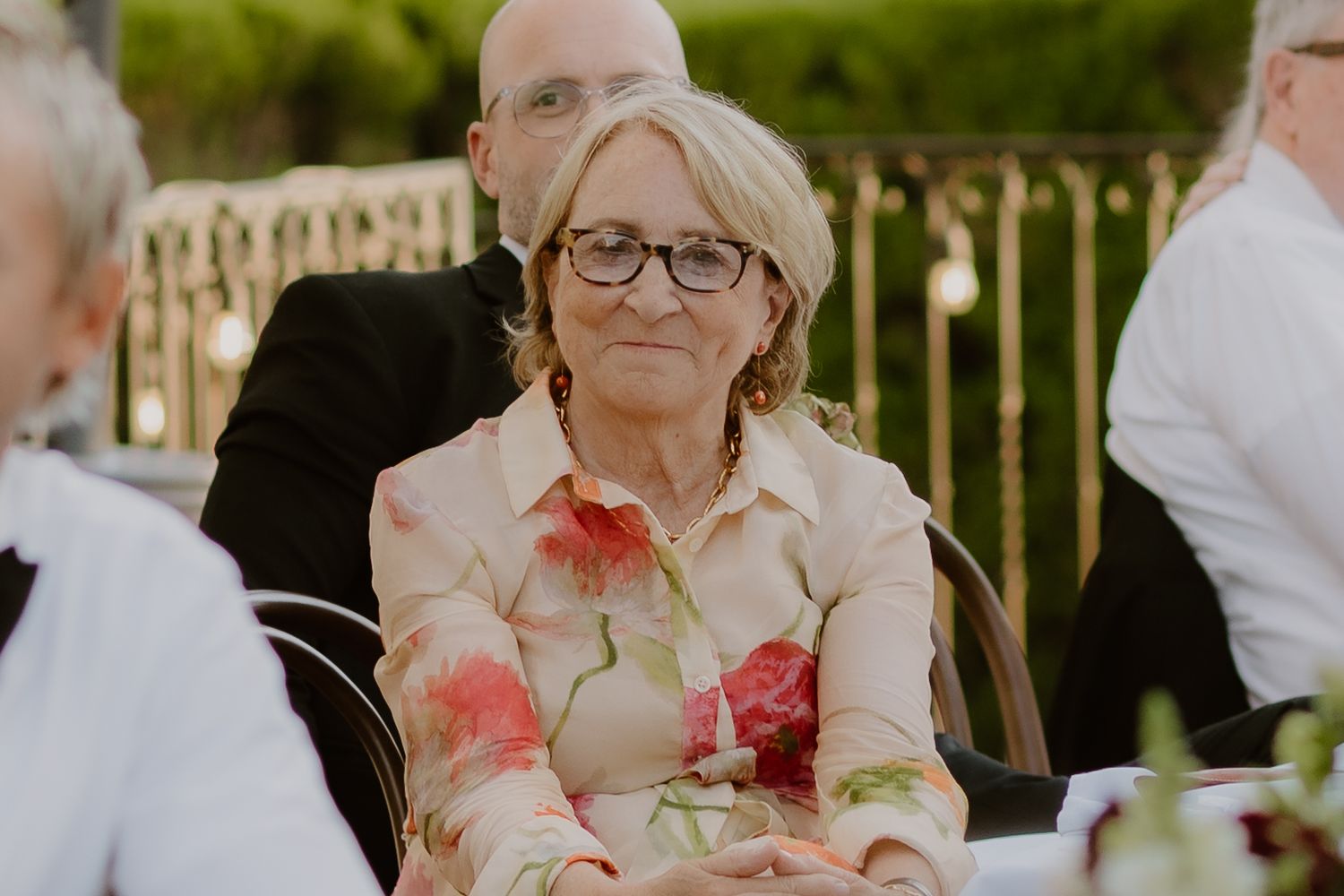 Elegant older woman in a floral blouse sits at an outdoor wedding reception table.