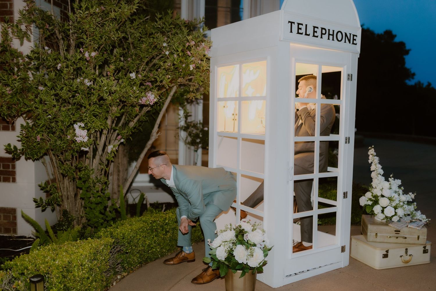 Wedding guest peeks into illuminated white telephone booth photo booth at evening reception.