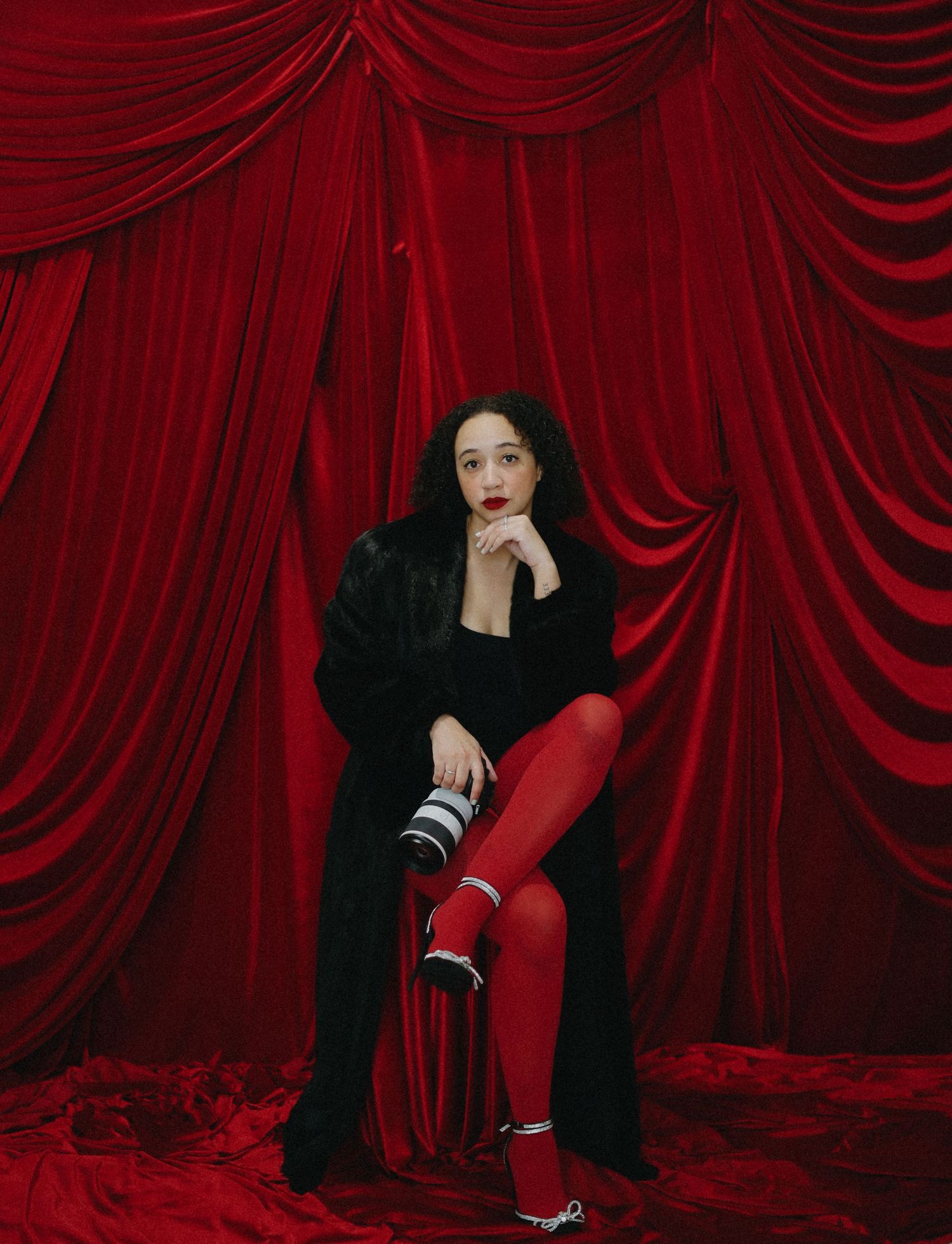 Woman in black fur coat and red tights sits elegantly before red velvet drapes.
