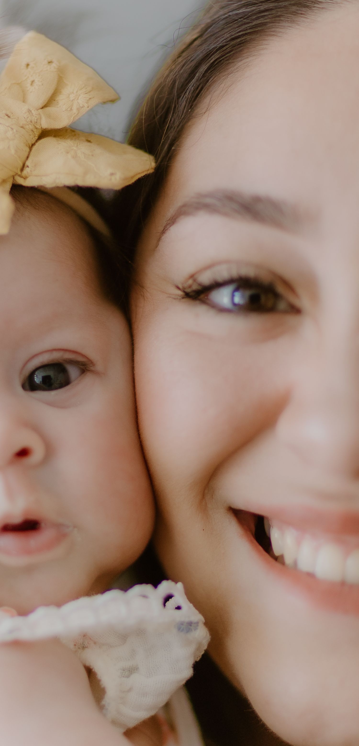 Close-up color portrait of a smiling mother and baby girl with a yellow bow headband pressed cheek-to-cheek.