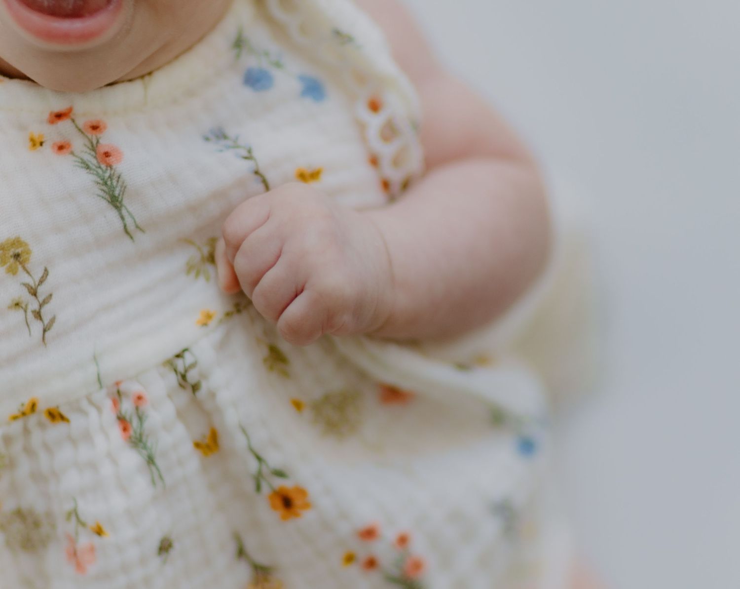 Close-up of baby girl in cream floral muslin dress with tiny chubby hands.