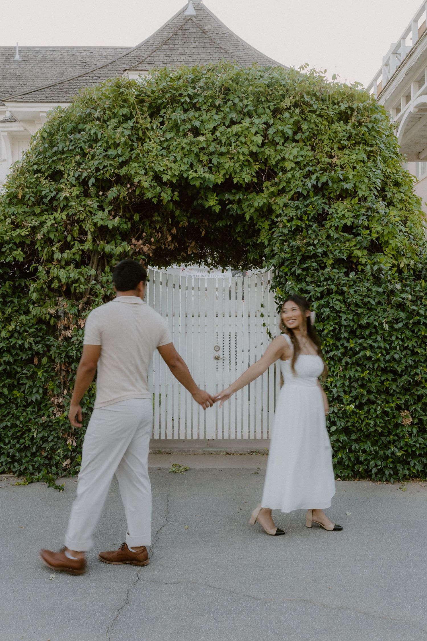 Couple holding hands walking toward ivy-covered white picket gate entrance together.