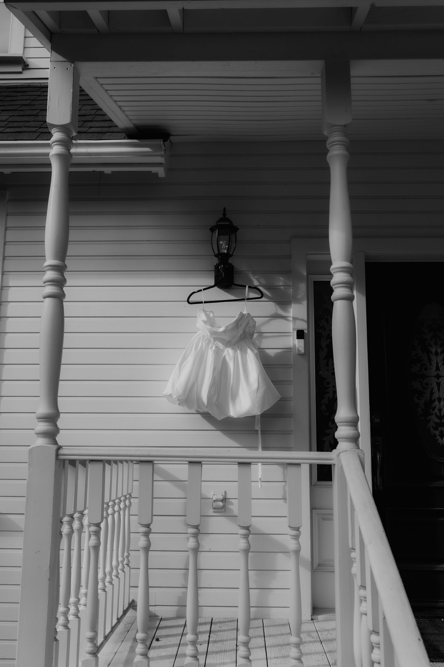 Black and white photo of white flower girl dress hanging on porch lantern of classic Victorian home.