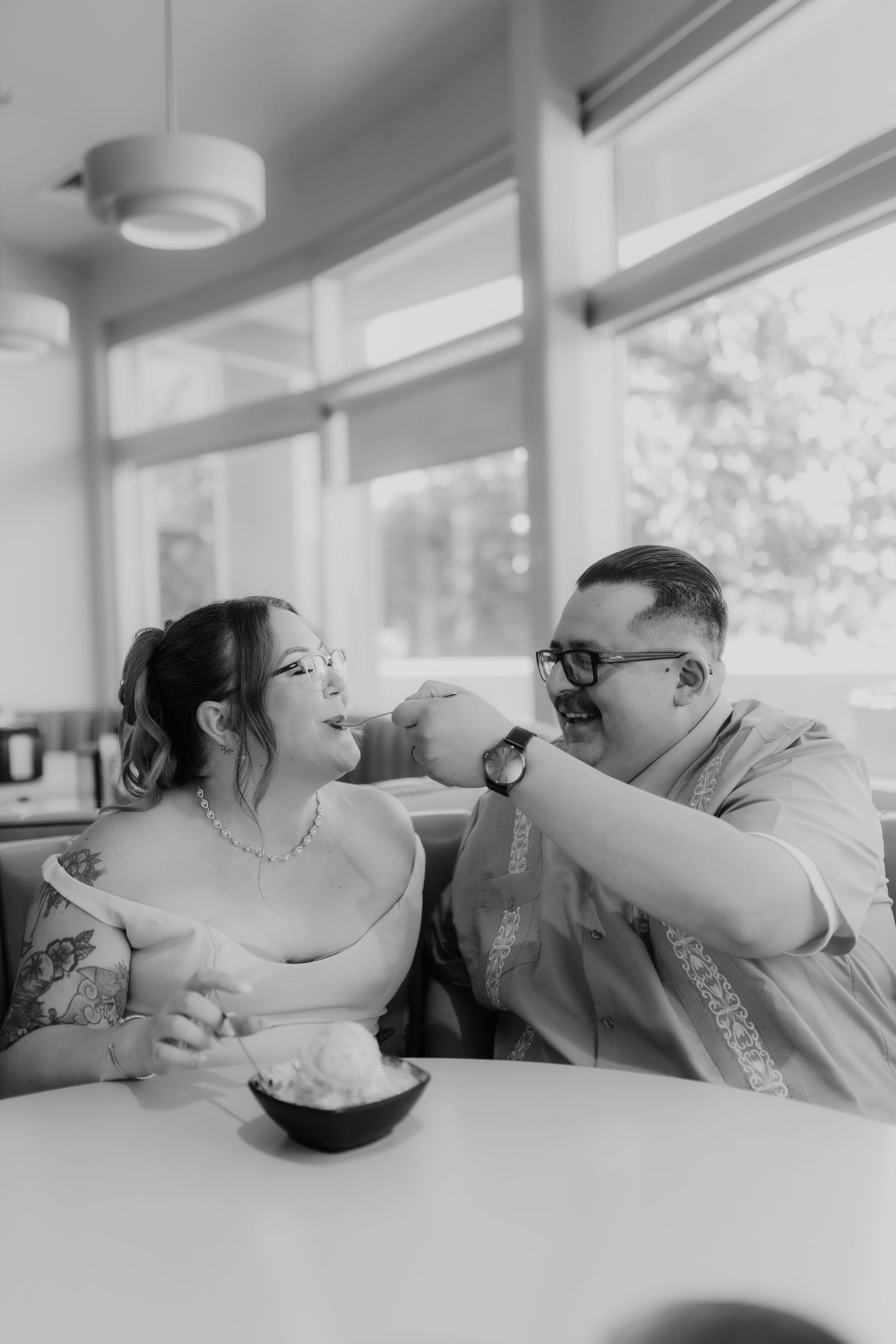 Black and white photo of couple playfully sharing ice cream at bright retro diner booth table.