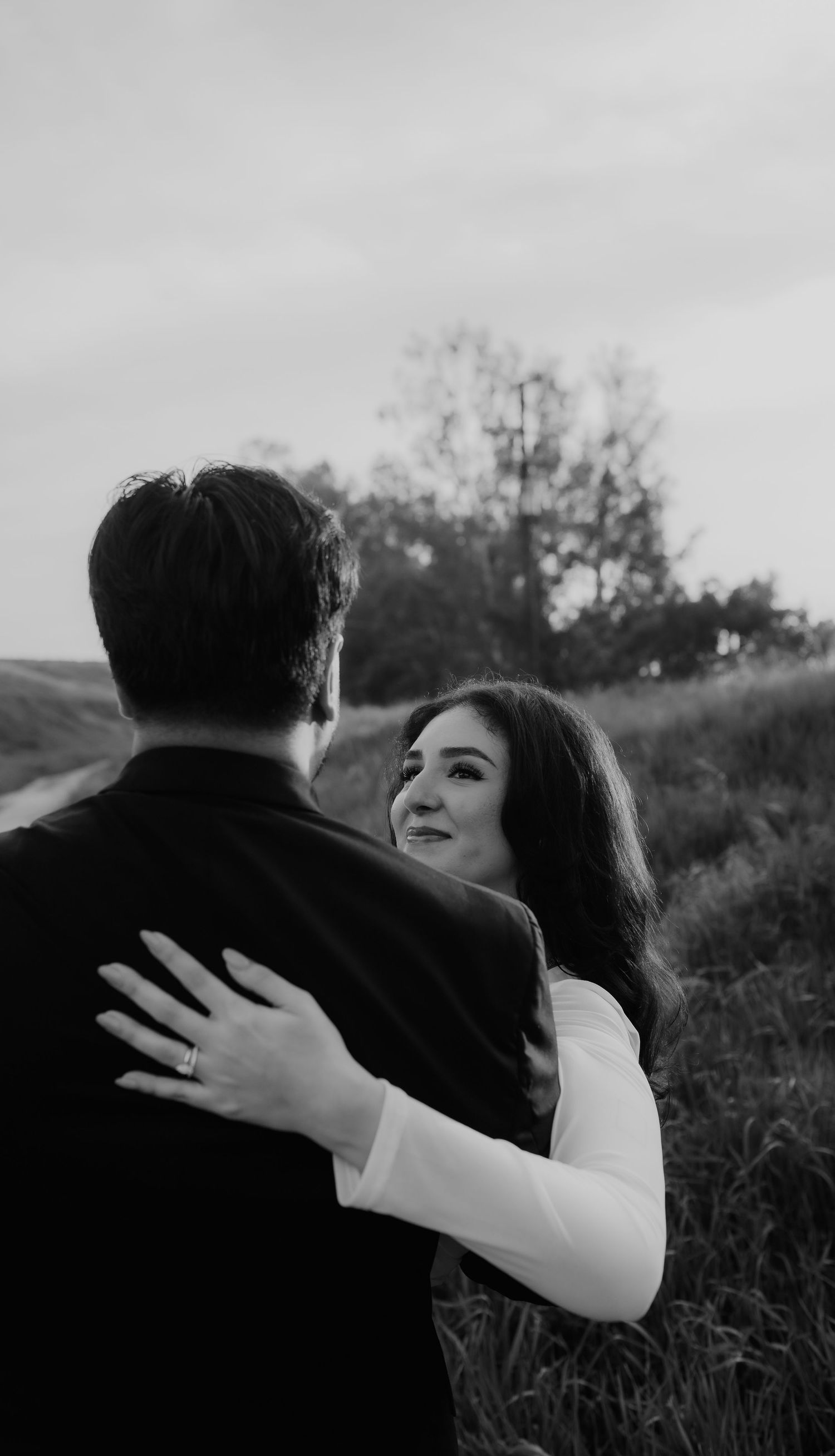 Black and white photo of a woman gazing at her partner while embracing outdoors in a field.