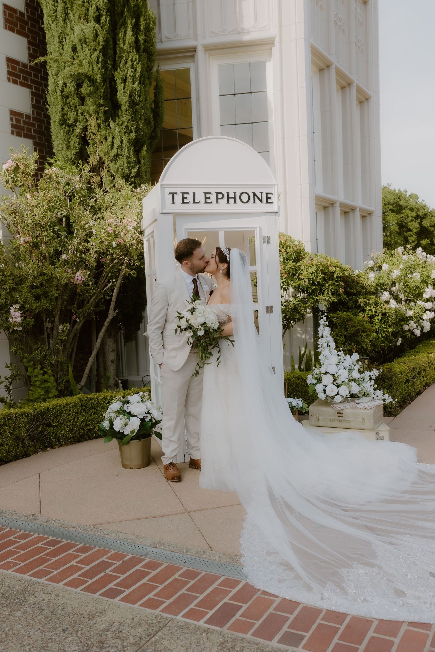 Bride and groom share a kiss inside a white telephone booth surrounded by lush garden florals.