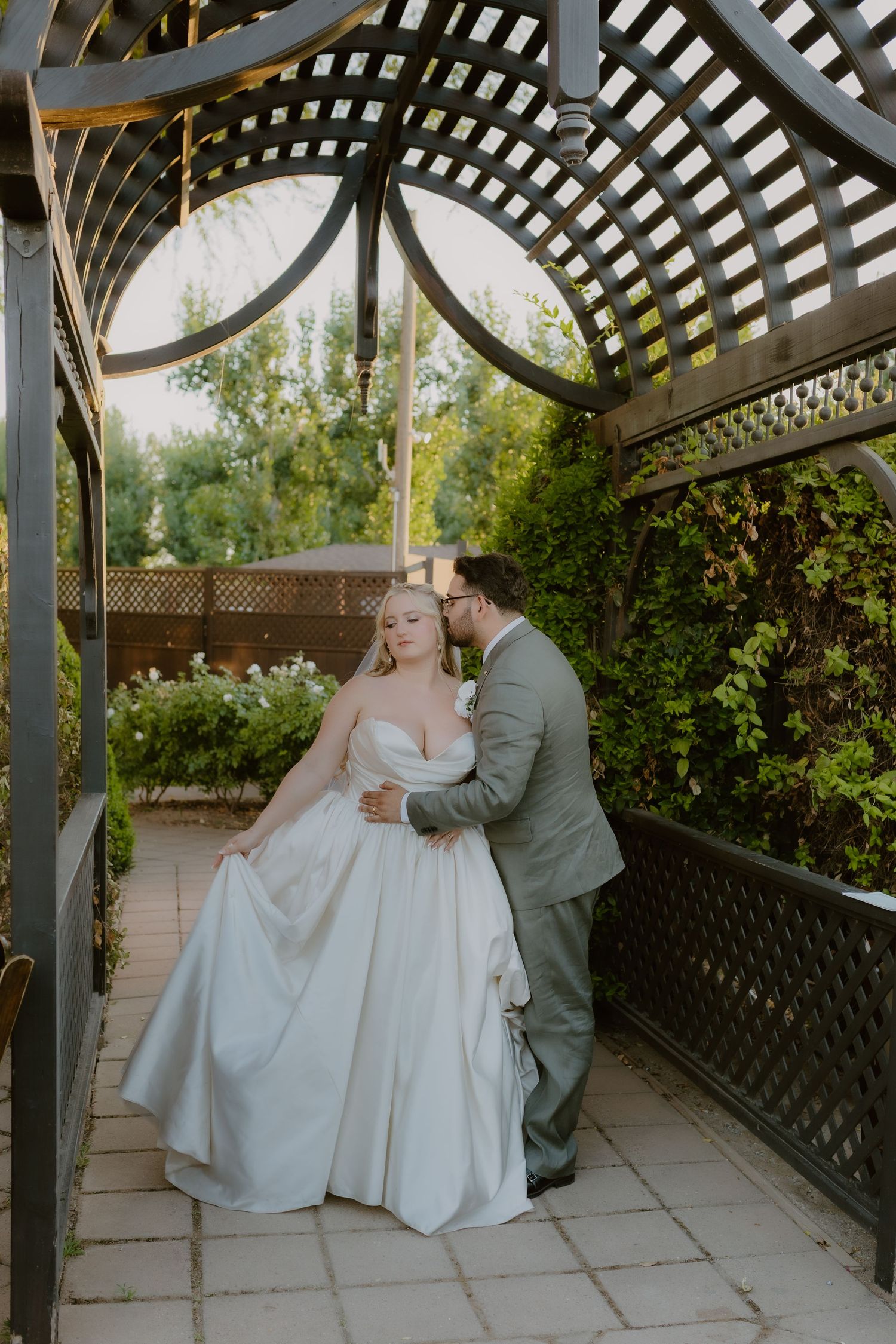 Bride and groom share a romantic moment under a wooden pergola surrounded by lush greenery at an outdoor wedding.
