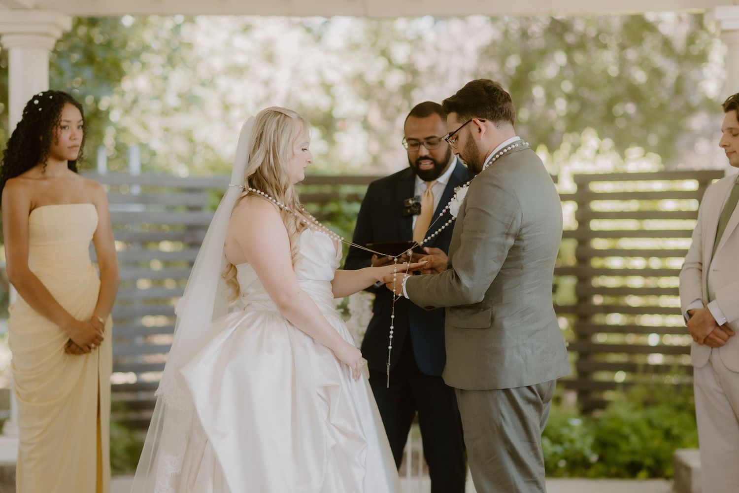 Bride and groom exchange rings during an outdoor wedding ceremony with bridesmaids and officiant present.