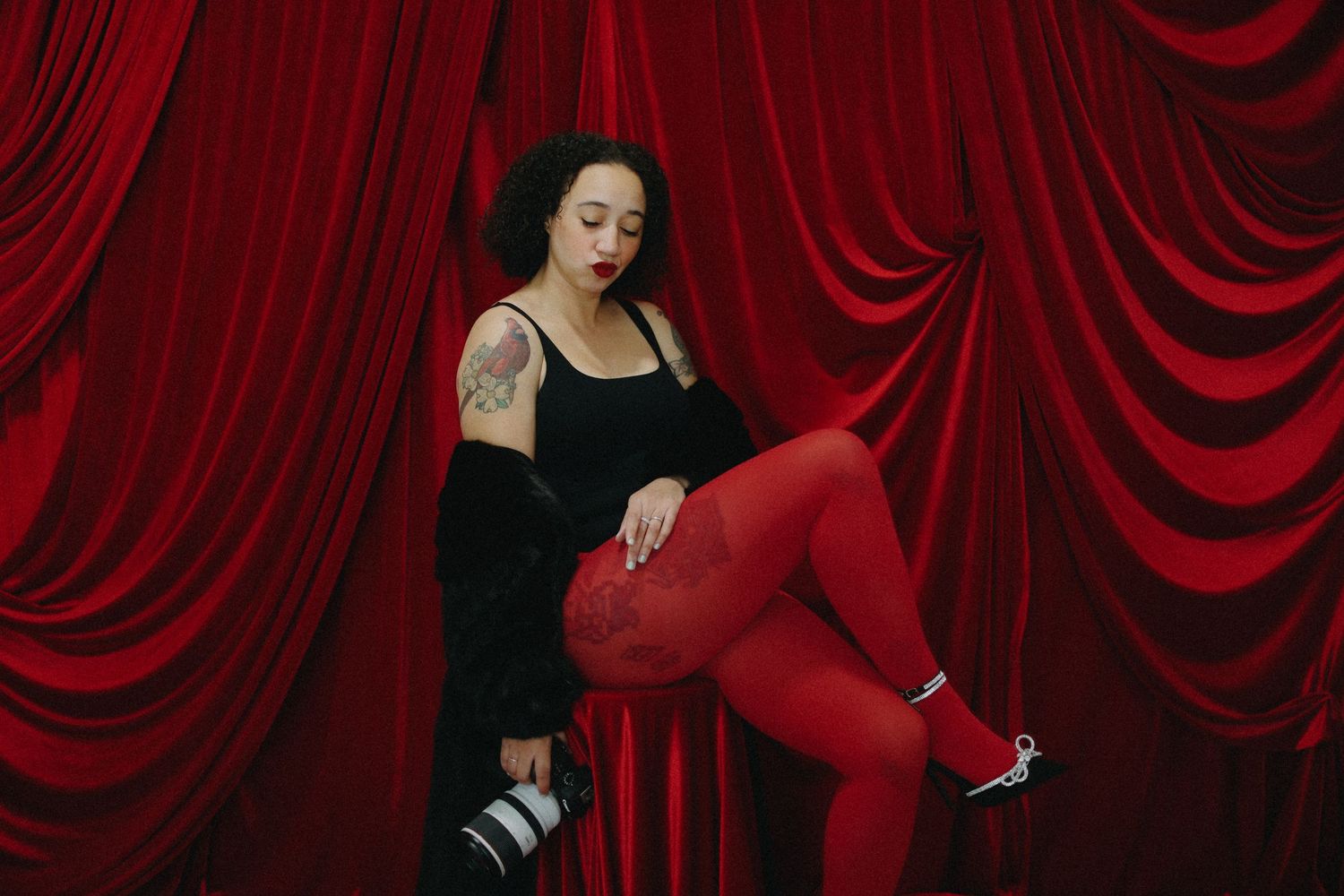 Woman in red tights and black top poses with camera against red velvet curtains.
