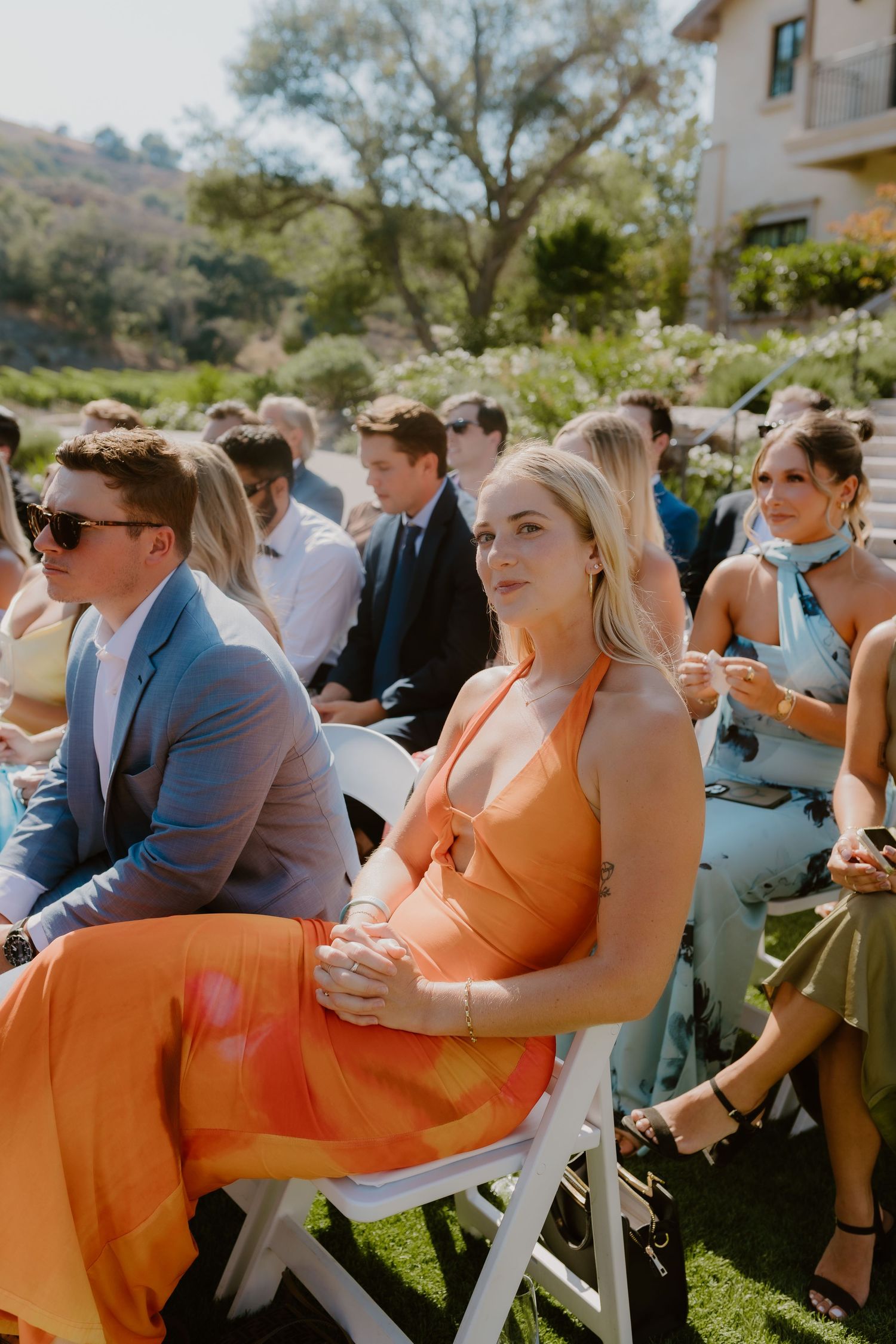 Wedding guest in vibrant orange halter dress seated at an outdoor garden ceremony.