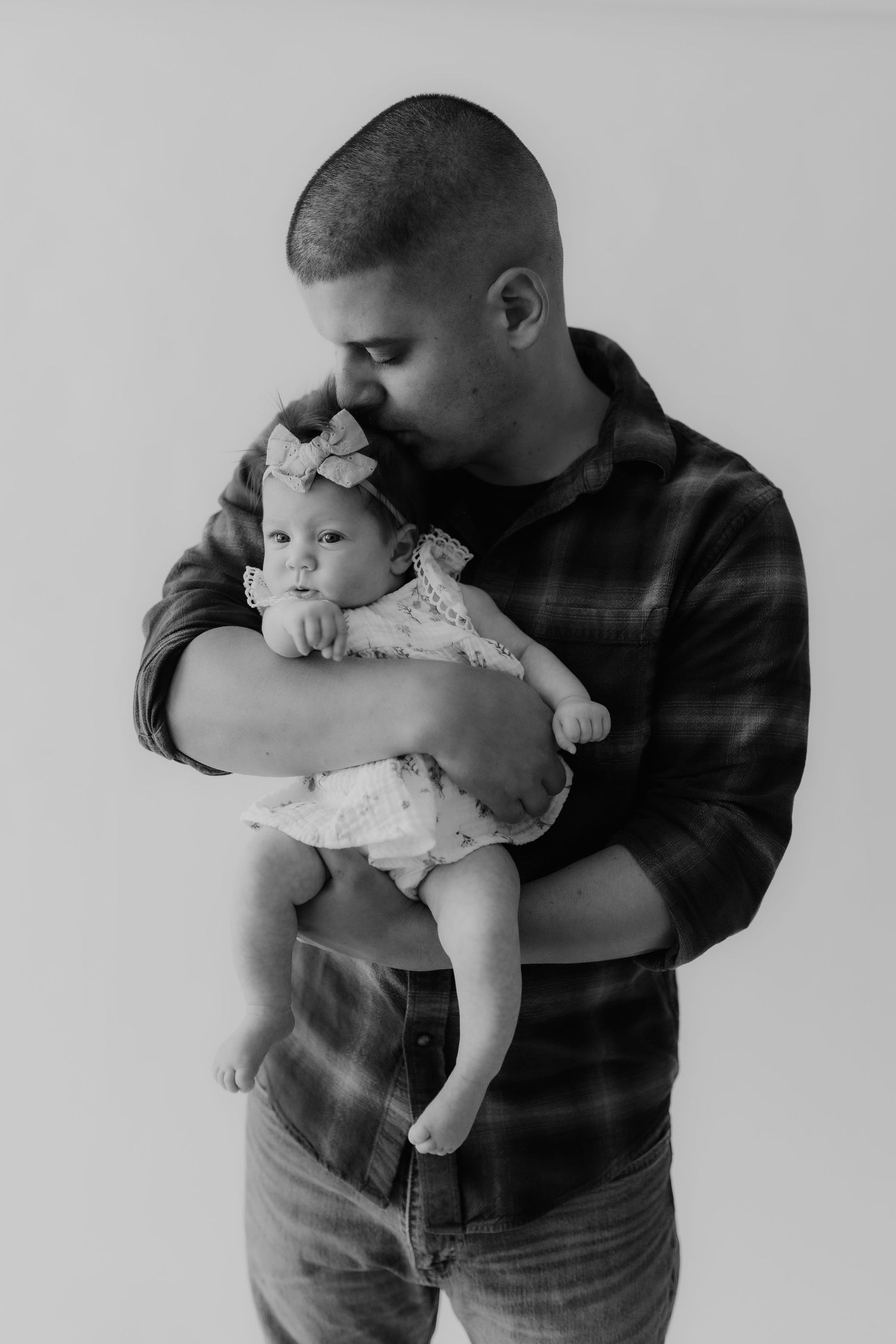 Black and white portrait of a father kissing his infant daughter wearing a bow headband and lace outfit.