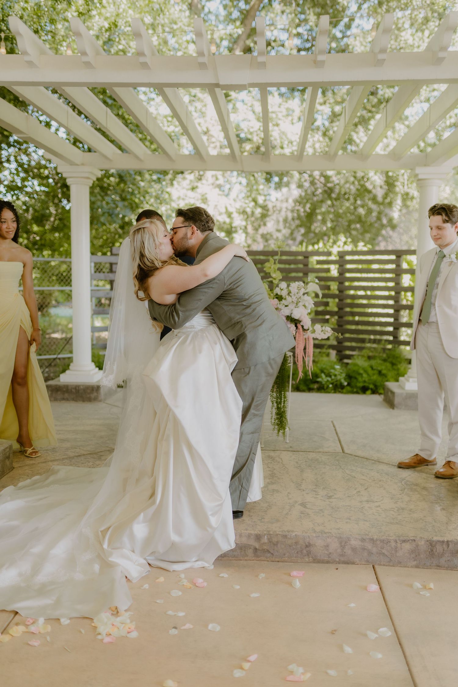 Newlyweds sharing first kiss embrace under white pergola at outdoor garden wedding ceremony.
